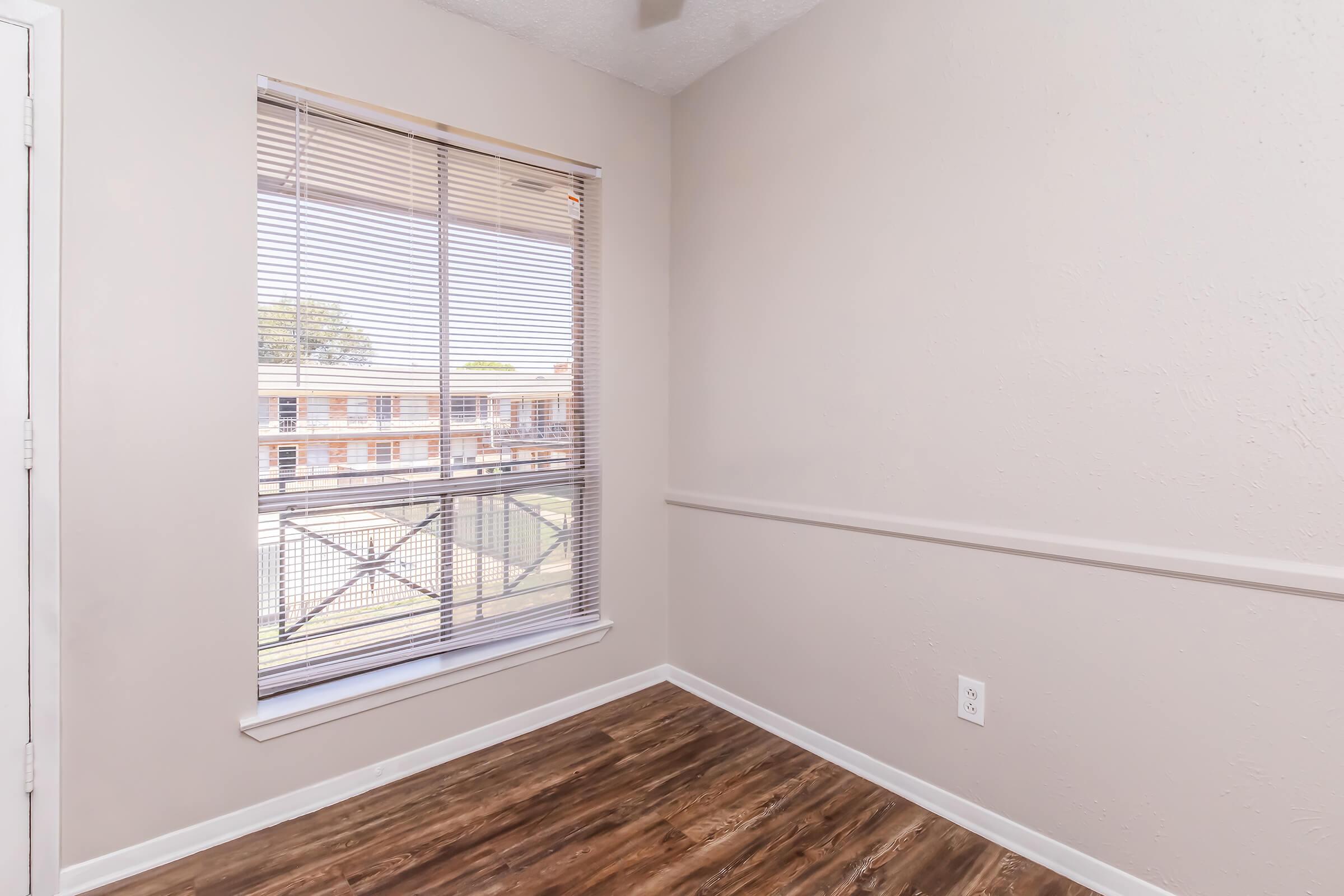 A small, empty room with beige walls and a wood laminate floor. A window with blinds brings in natural light, overlooking an exterior balcony and nearby buildings. There are no furnishings, creating a minimalist and spacious appearance.