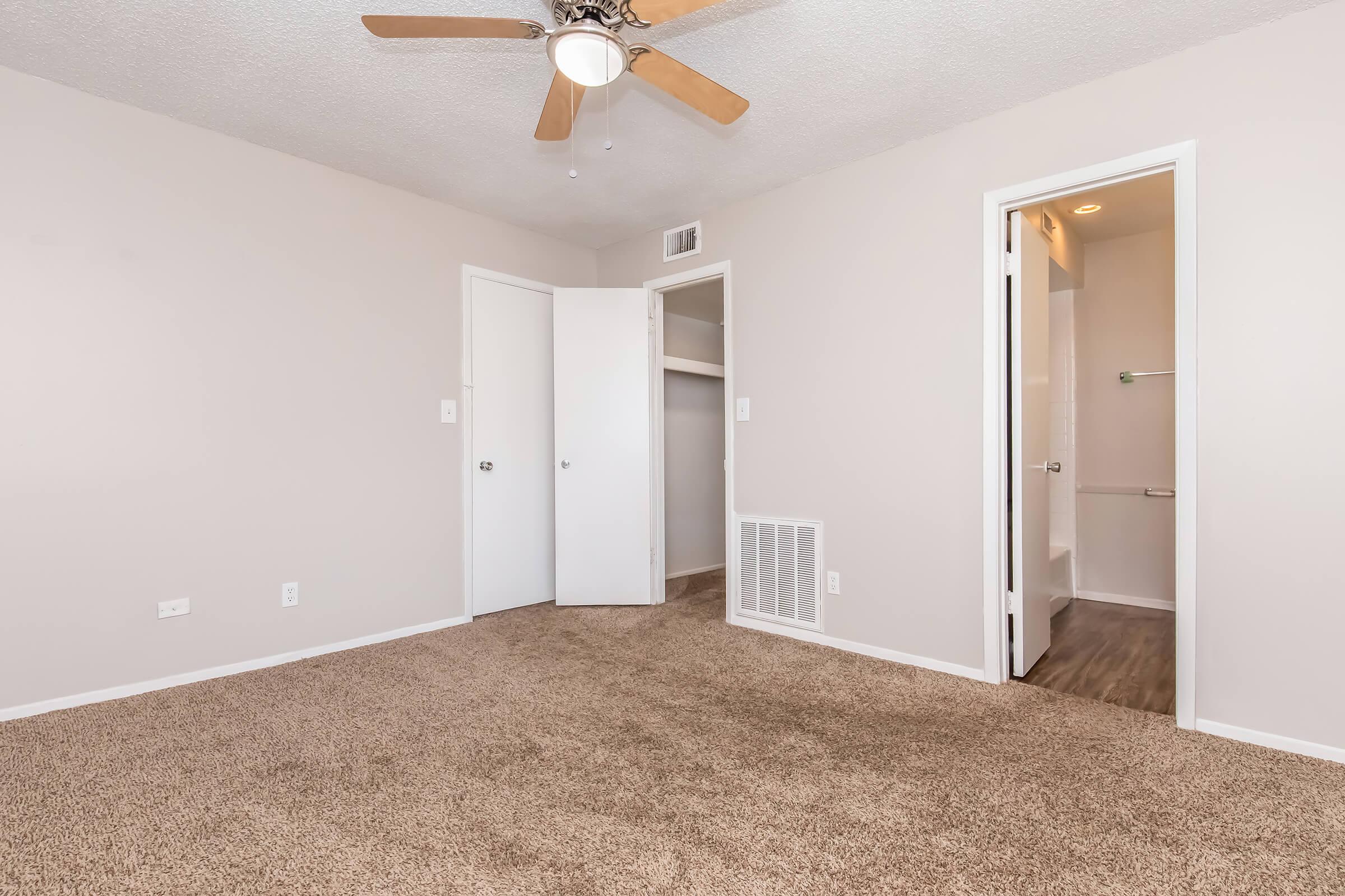 A spacious room with light beige walls and plush carpet. It features a ceiling fan, a closet with sliding doors, and a doorway leading to a bathroom. The room is well-lit, showcasing a clean and modern aesthetic, ideal for a comfortable living space.