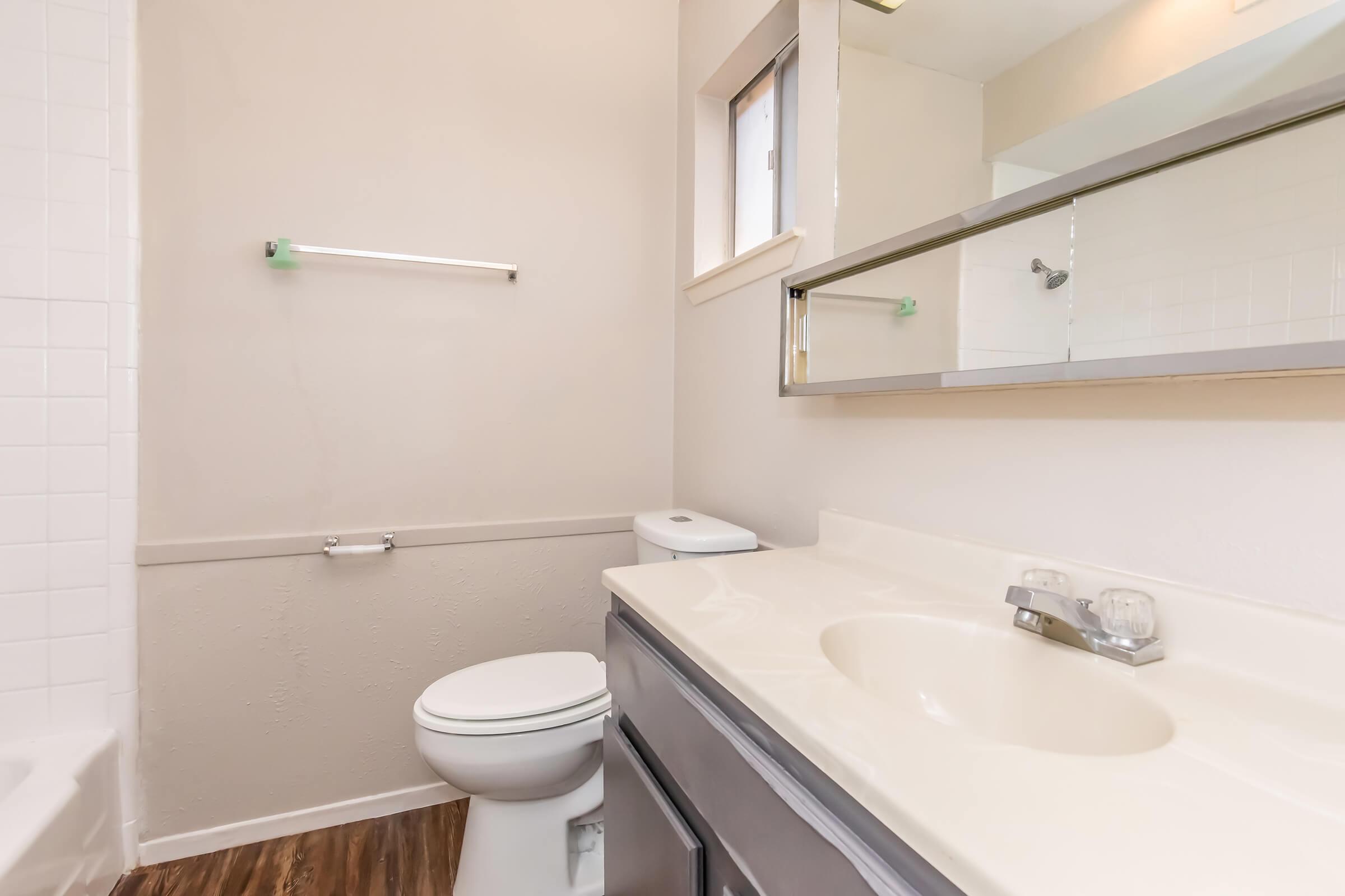 A clean, modern bathroom featuring a white toilet, a light-colored vanity with a sink, a large mirror above the sink, and a wall-mounted towel rack. The walls are painted in a neutral tone, and there is a window allowing natural light. The flooring is a wood-like laminate.