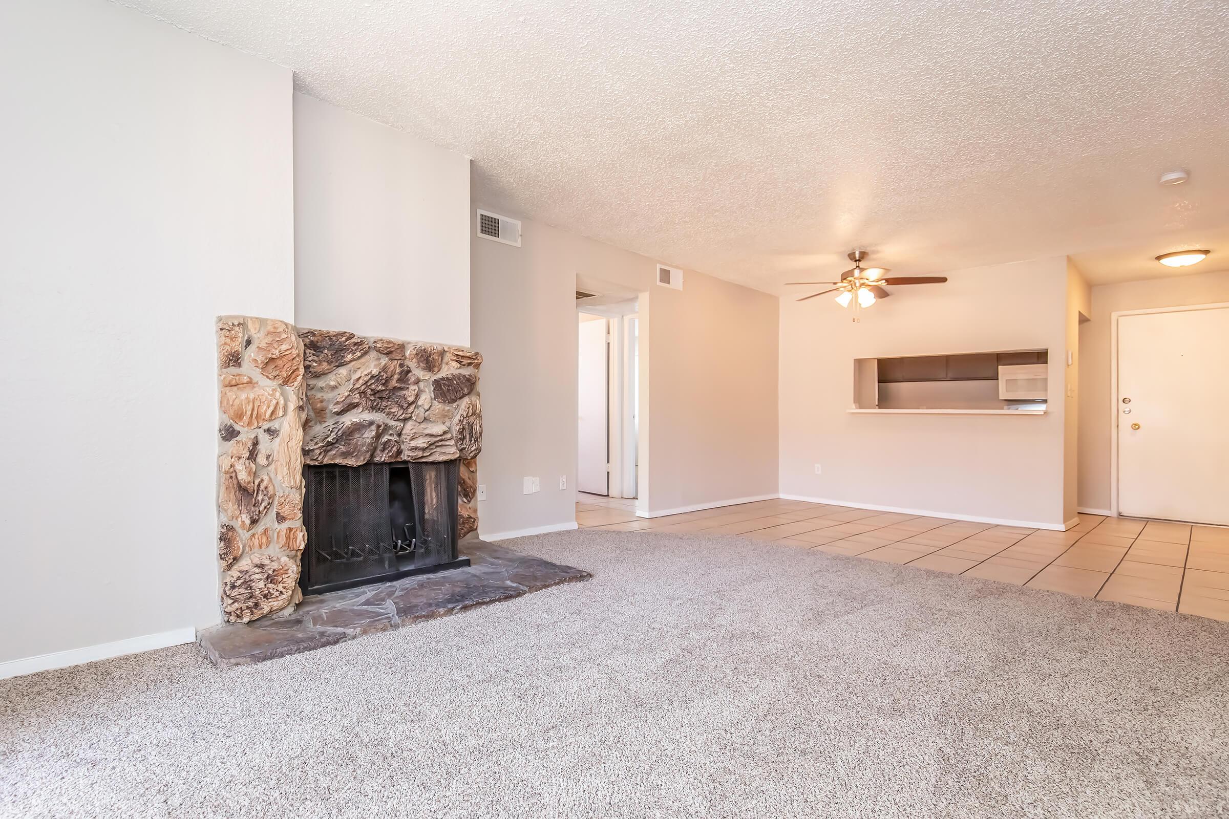 Spacious living room featuring a stone fireplace, plush carpet, and ceiling fan. The room opens to a kitchen area with tile flooring, and there is a doorway leading to the rest of the home. Soft, neutral colors create a warm and inviting atmosphere.