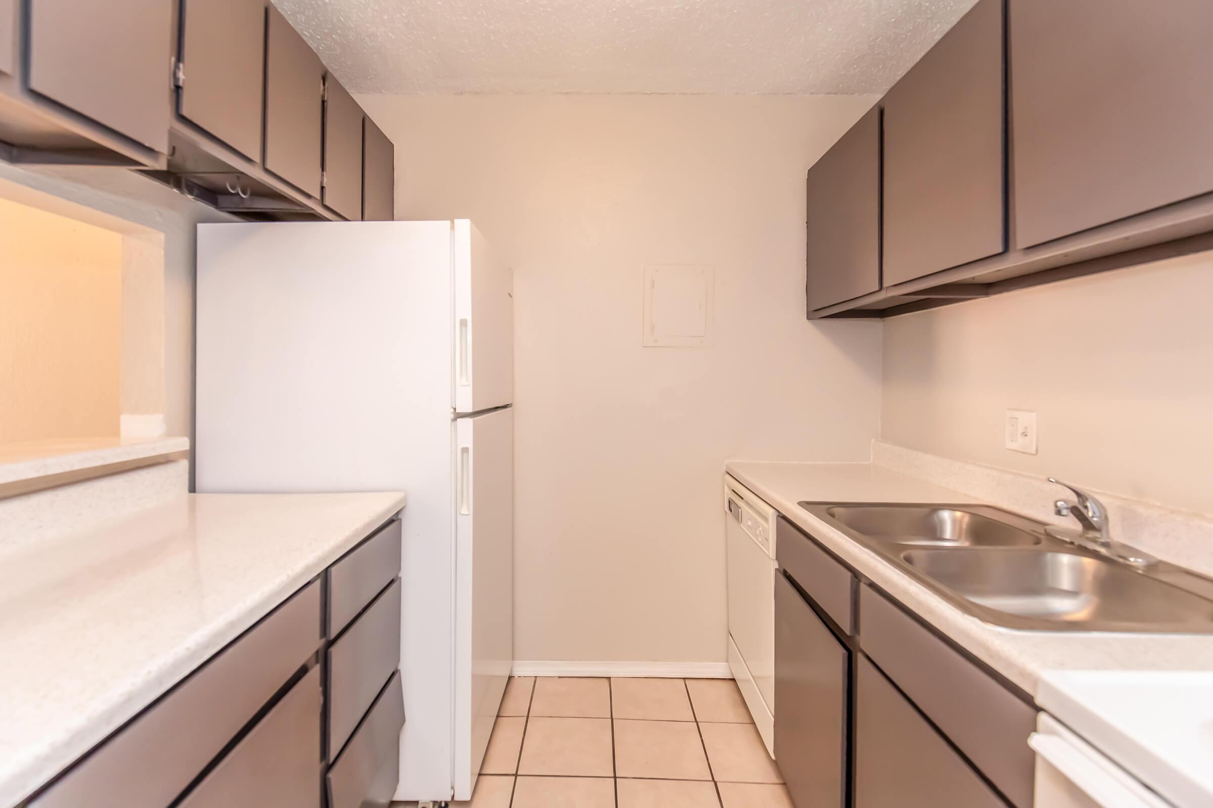 A small kitchen featuring dark cabinetry, a white refrigerator, and a dishwasher. The countertops are light-colored, and there's a double sink. The floor is tiled, and the walls are a neutral shade, creating a bright and clean space.