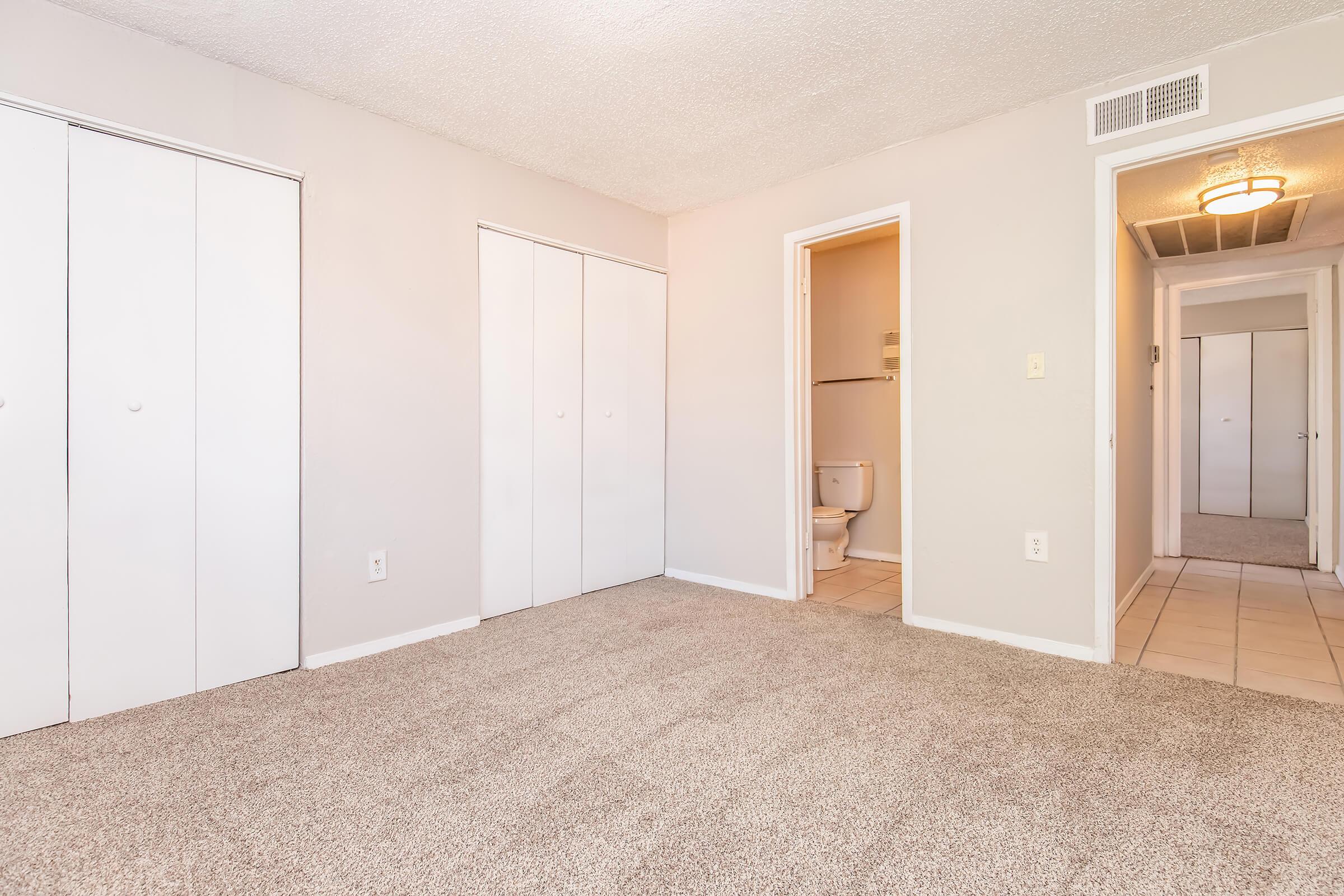 A spacious room with light gray walls and plush carpet. Two closed white closet doors are visible on the left. A doorway leads to a bathroom on the right, featuring a toilet. A hallway can be seen in the background, with additional doors. Soft lighting from an overhead fixture adds warmth to the space.