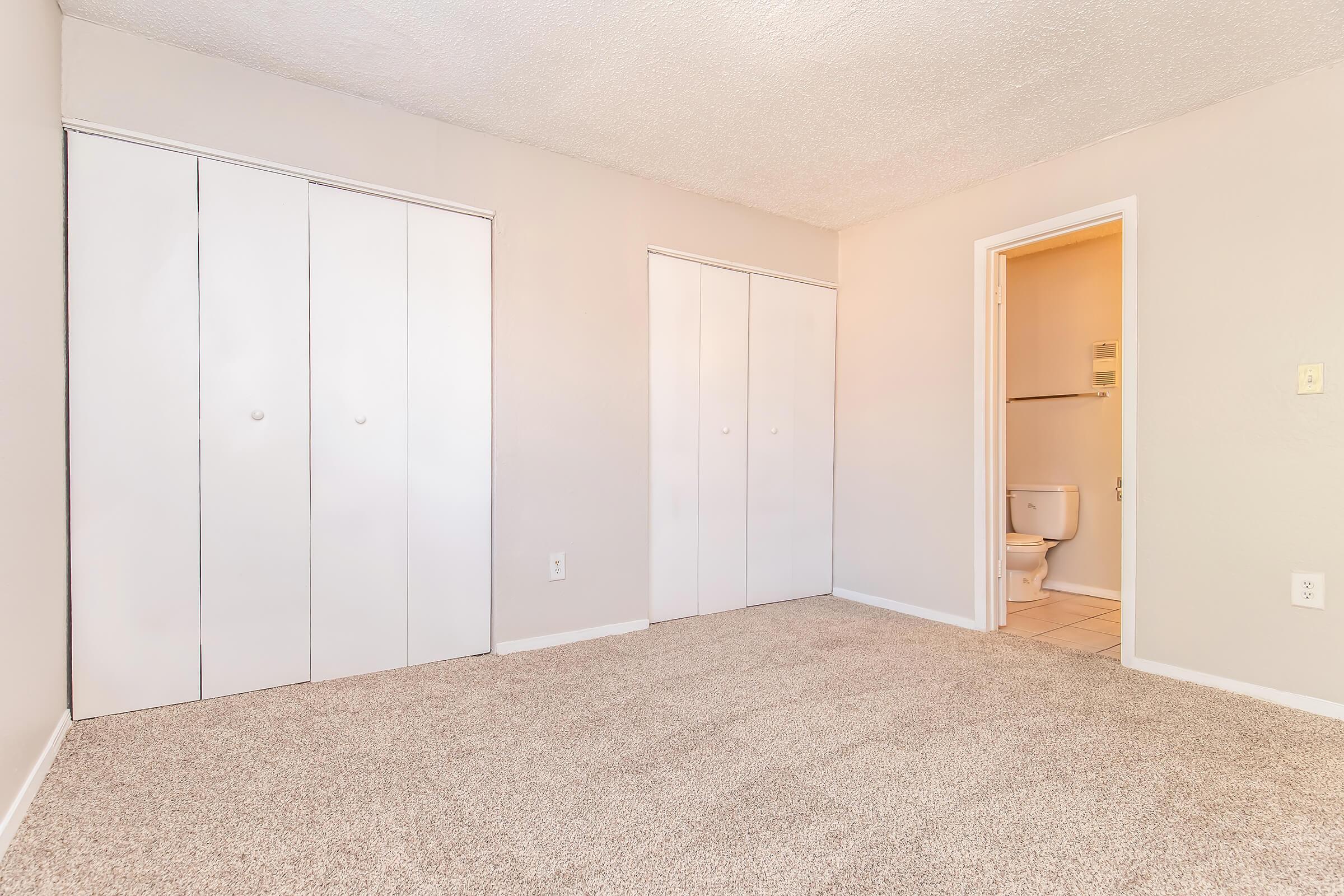 A clean, neutral-toned room with light gray walls and plush beige carpet. The room features two sets of white double doors for storage, opposite a bathroom door which is partially visible. Natural light brightens the space, creating a calm and inviting atmosphere.