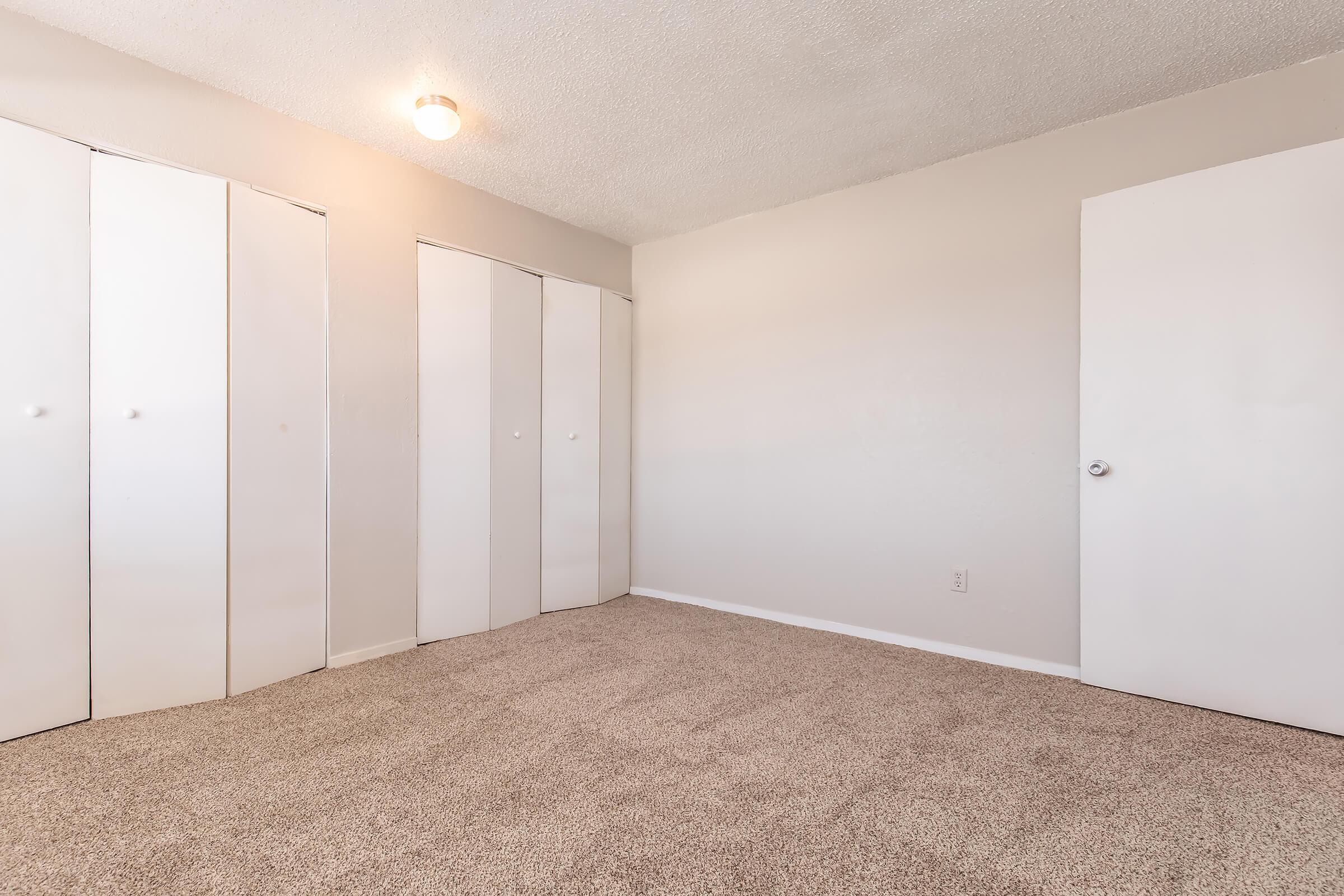 A spacious, empty bedroom featuring light gray walls, soft carpet flooring, and a ceiling light fixture. There are three white sliding closet doors on the left and a white door leading out of the room on the right, creating a simple and clean aesthetic. Ideal for personalizing with furniture and decor.