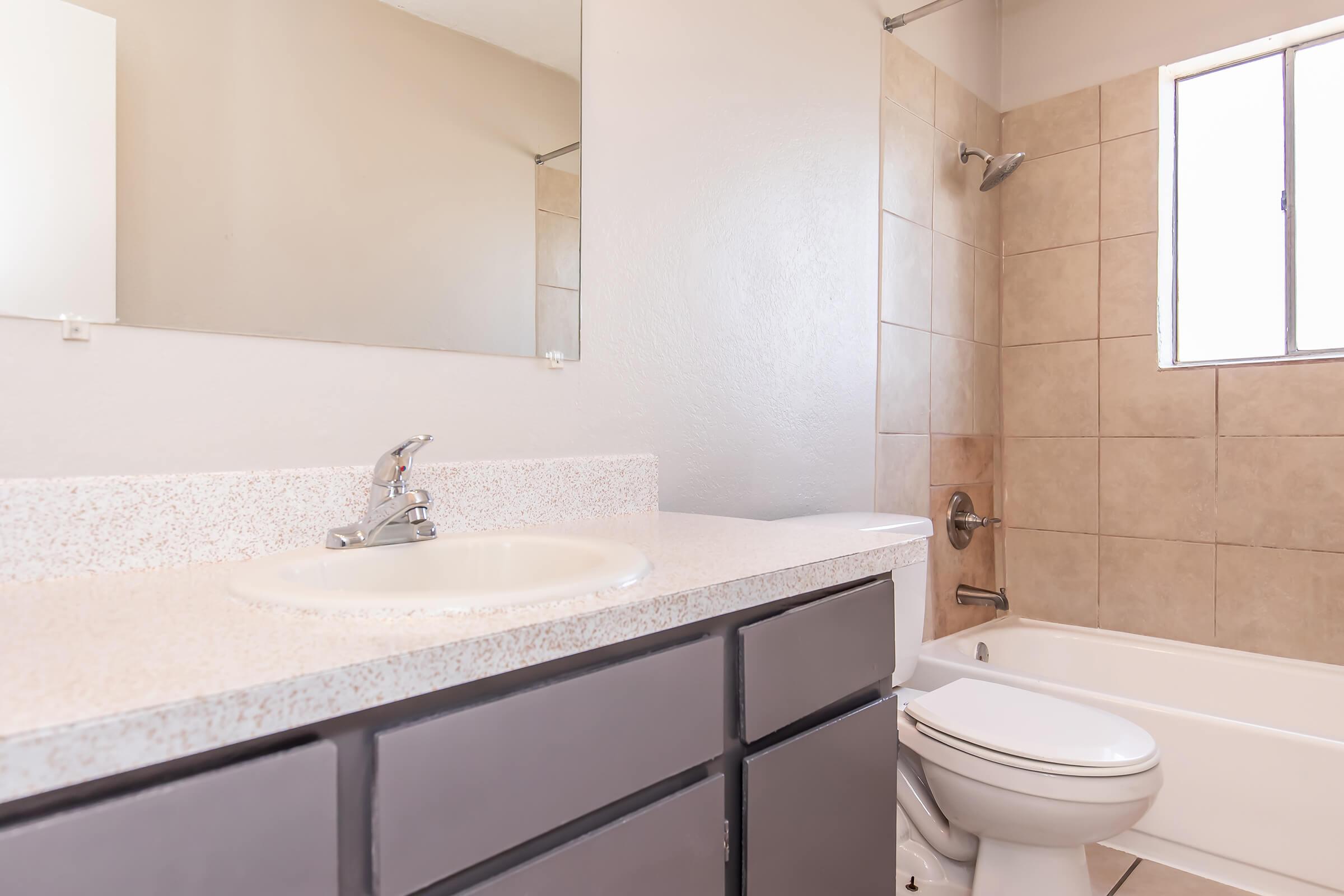 A clean, modern bathroom featuring light-colored tiled walls, a bathtub with a shower, a white toilet, and a dark gray vanity with a sink. A mirror hangs above the sink, and natural light enters through a window, creating a bright and airy atmosphere.