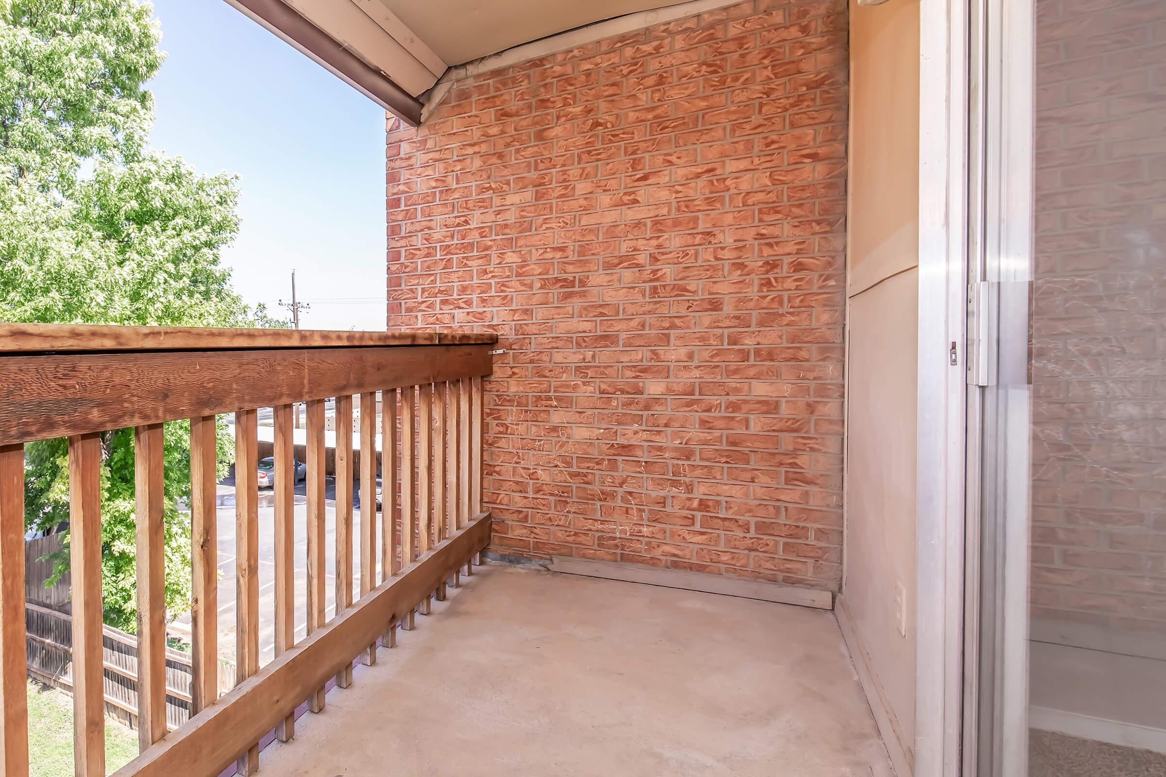 A small balcony with a wooden railing, featuring a brick wall on one side. The floor is concrete, and there are trees visible outside, suggesting a sunny day. The scene is simple and uncluttered, providing a cozy outdoor space.