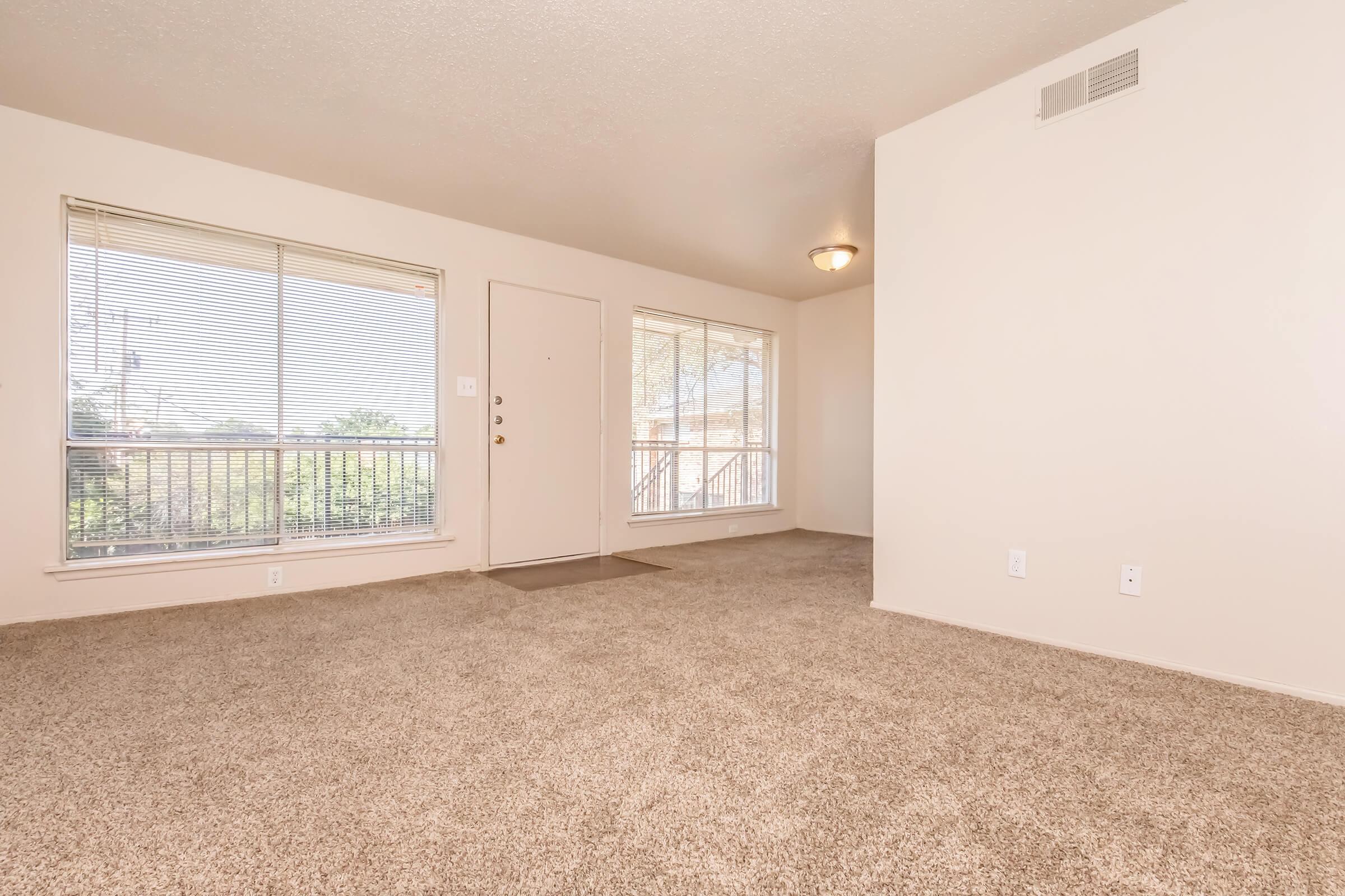 Empty living room with light beige walls and carpet. There are two large windows allowing natural light, a front door, and a light fixture on the ceiling. The space is uncluttered and showcases a welcoming atmosphere.