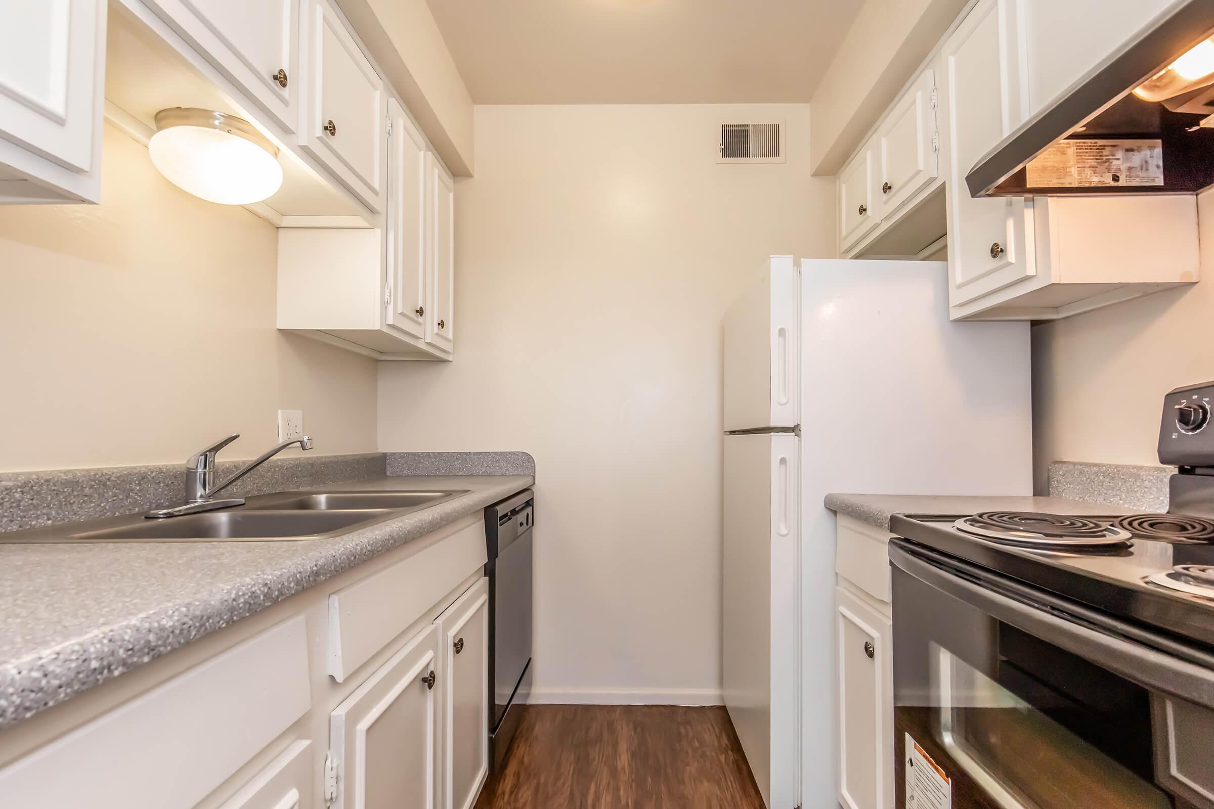 A small kitchen featuring white cabinets, a stainless steel sink with a double basin, a dishwasher, a white refrigerator, and a stove with an oven. The countertops are gray, and the walls are painted in a light color, creating a bright and clean appearance.