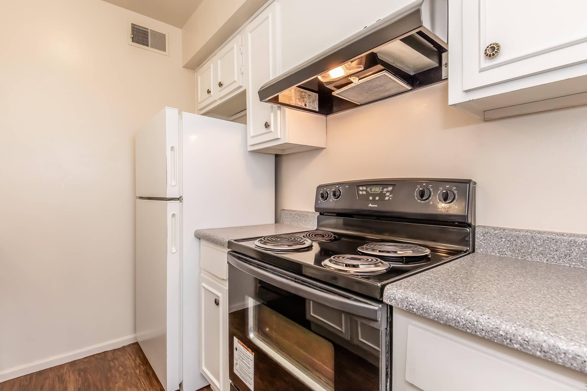 A clean and modern kitchen featuring a white refrigerator, a black electric stove with an oven, and white cabinetry. The countertops are gray, and there is a ventilation hood above the stove. The walls are light-colored, contributing to a bright and spacious atmosphere.