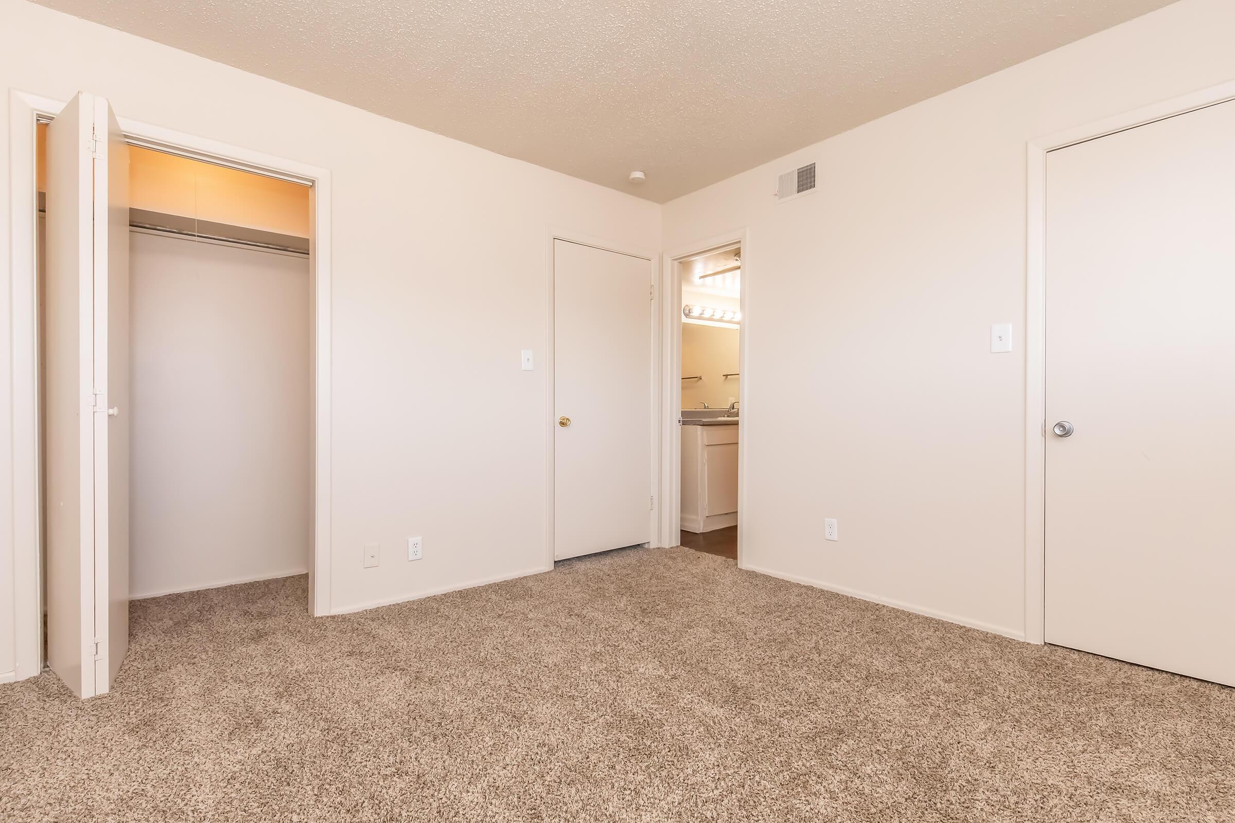 A clean, empty bedroom with light beige walls and plush, beige carpet. It features two doorways: one leading to a closet and the other to a bathroom area. Natural light illuminates the space, creating a bright and inviting atmosphere. The room is well-maintained and ready for decoration.