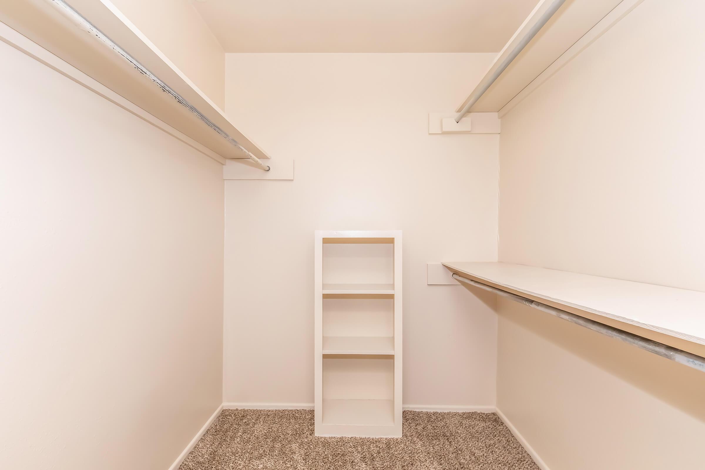 A small, empty closet featuring white walls and light-colored shelving. There is a central cabinet or shelving unit with three open compartments, and two horizontal shelves above are designed for hanging clothes. The floor is covered with tan carpet.