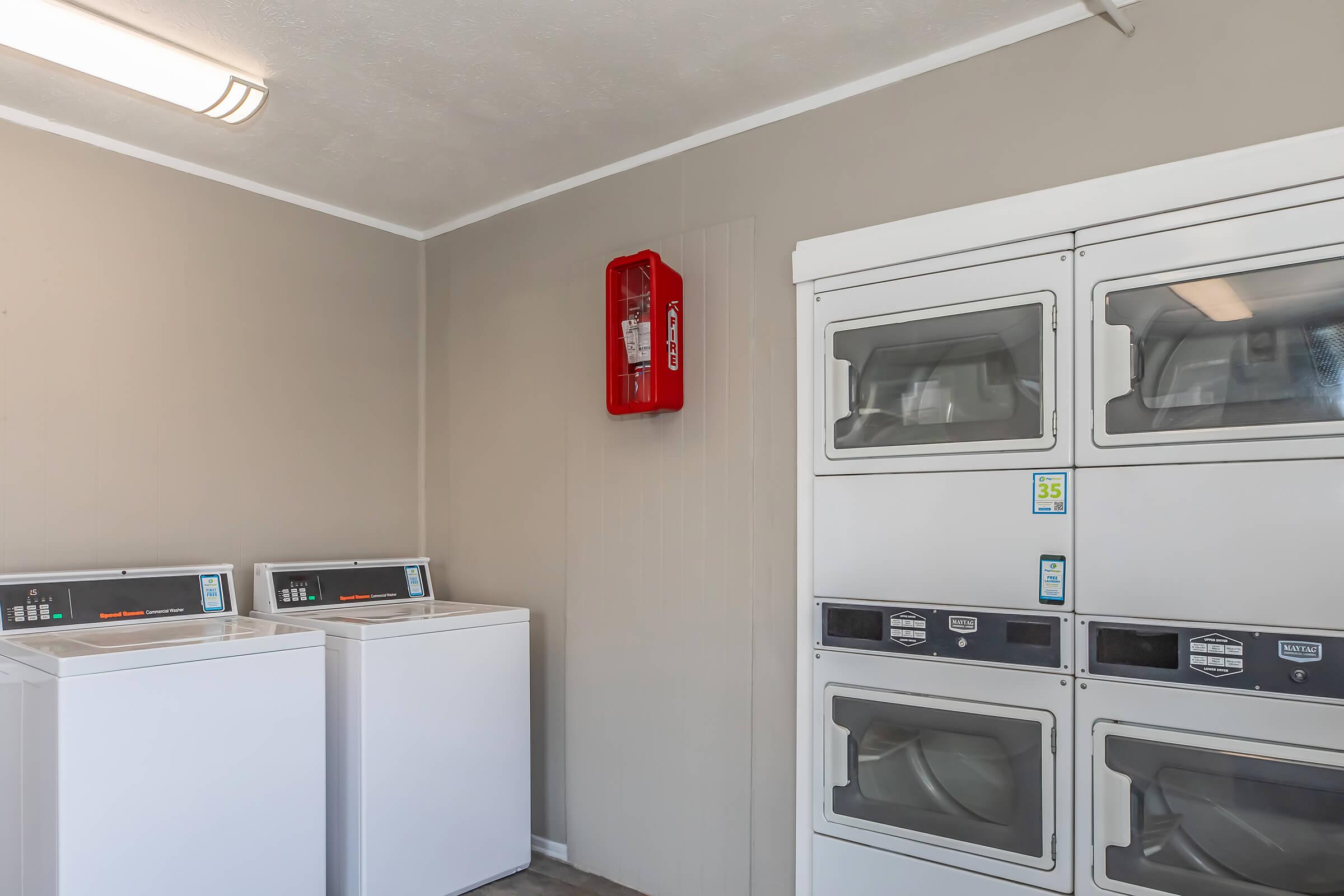 A clean laundry room featuring two white washing machines, a stack of coin-operated dryers, and a wall-mounted red fire extinguisher. The walls are painted a light gray, and there is bright overhead lighting illuminating the space.