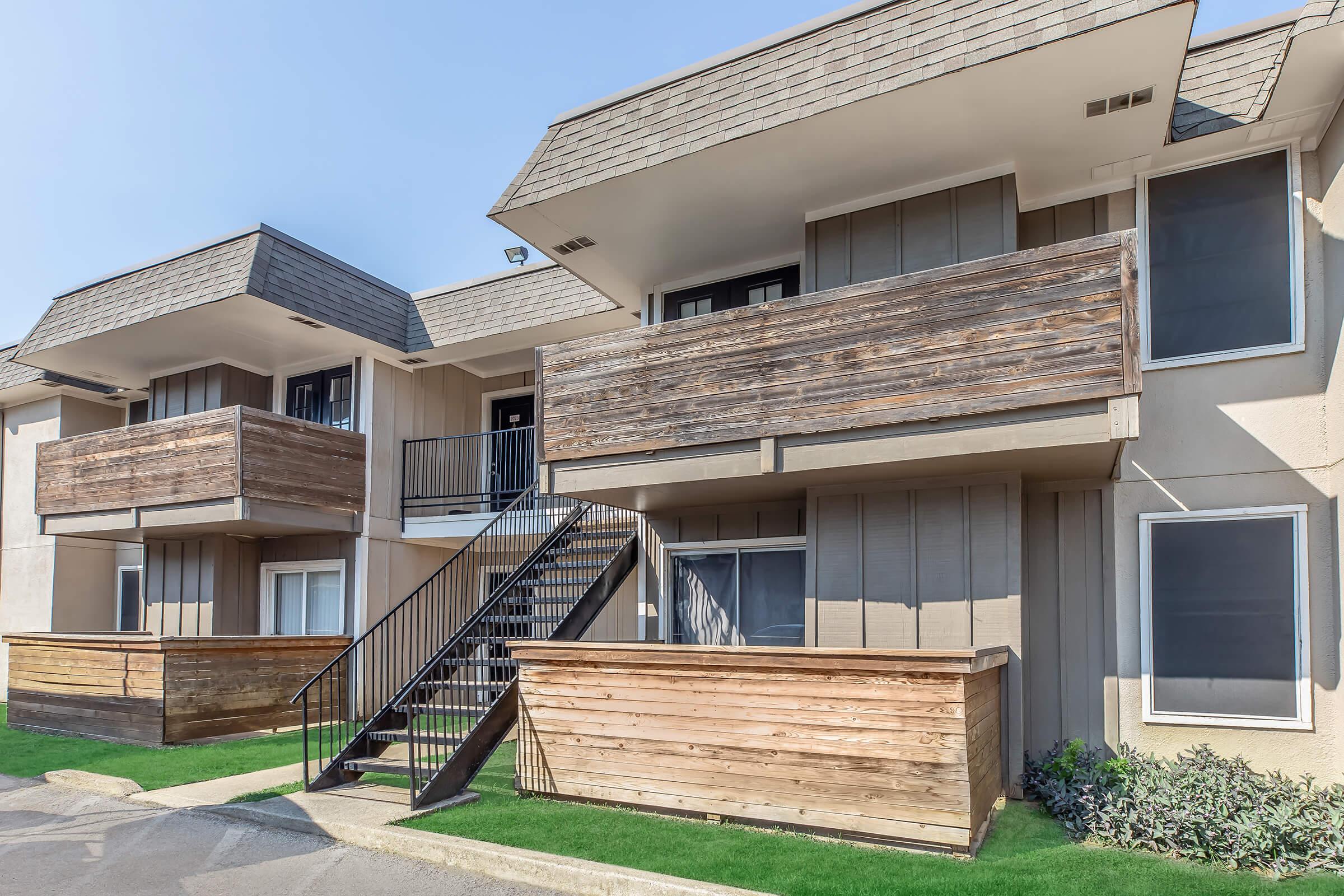 A view of two modern apartment buildings with wooden balconies and a staircase. The exterior features light-colored walls and wooden accents. The setting is bright with a clear blue sky, and there is greenery at the base of the buildings.