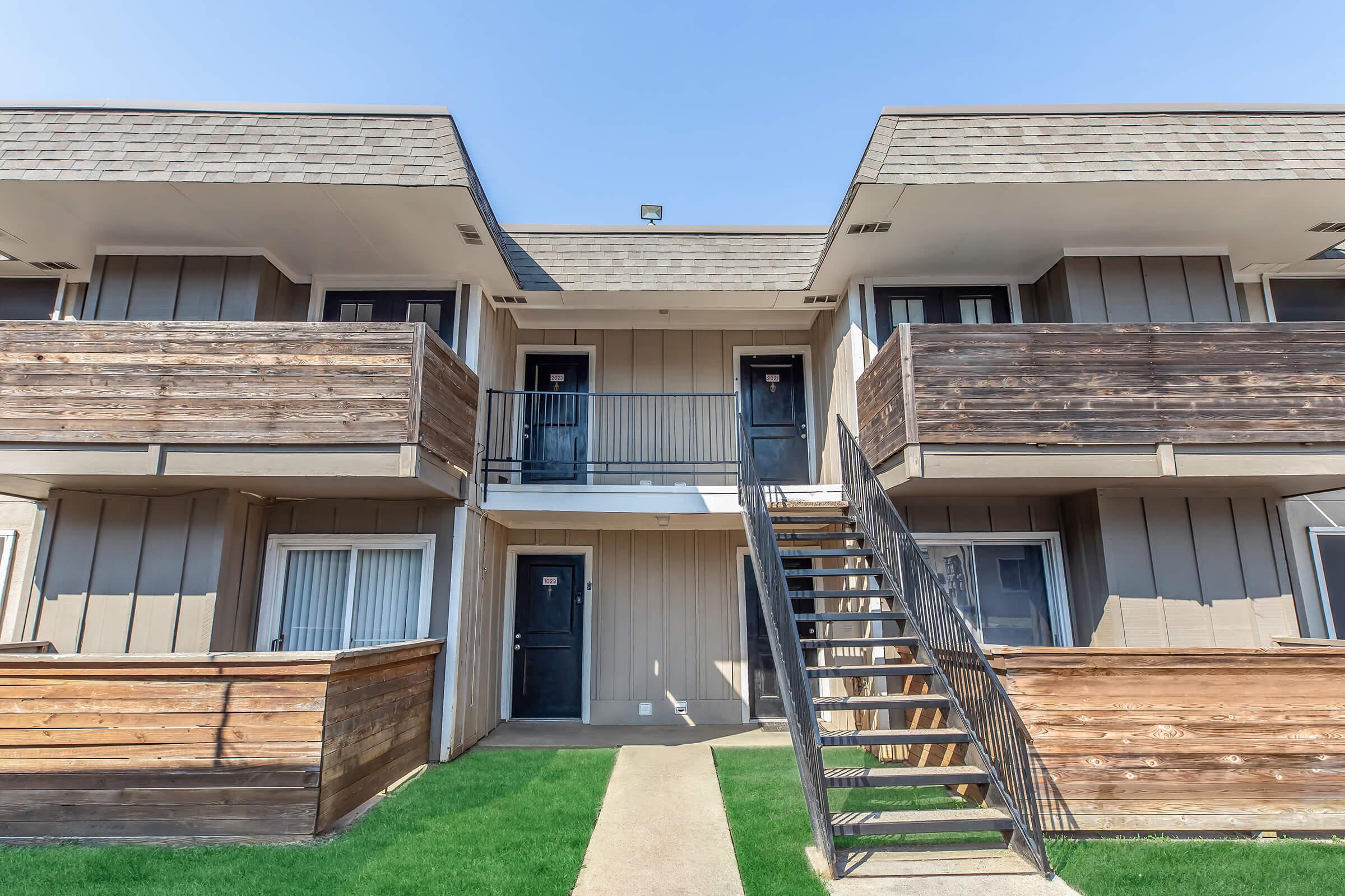 Two-story apartment building with a wooden exterior. Each floor has two units with black doors, and outdoor staircases provide access to the upper level. There are wooden railings and a small patch of green grass in the foreground. The sky above is clear and blue.