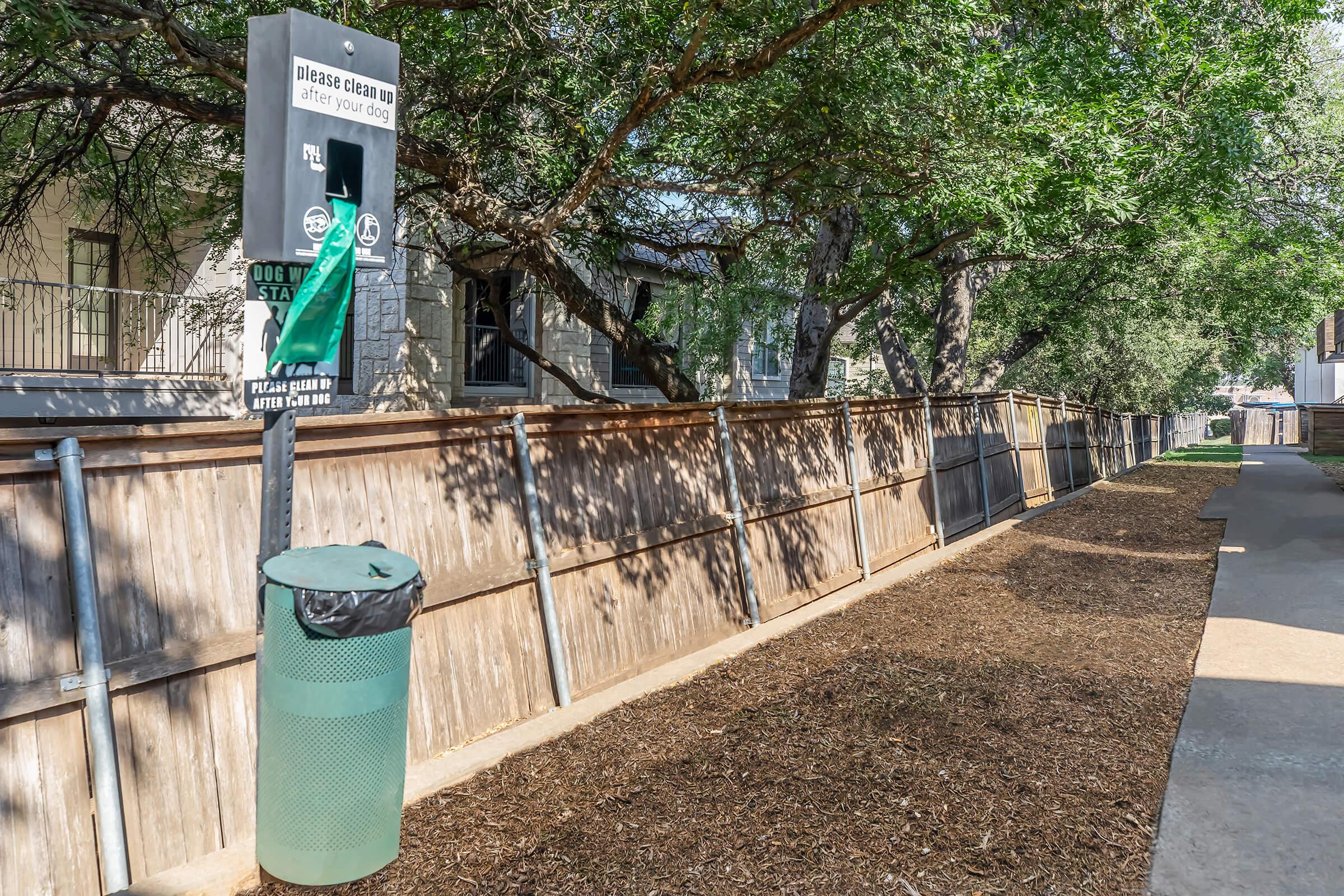 A dog waste station is set up along a gravel path surrounded by trees. The station includes a sign requesting dog owners to clean up after their pets, with a waste bag dispenser and a green trash bin nearby for disposal. The area appears well-maintained and is lined with a wooden fence.