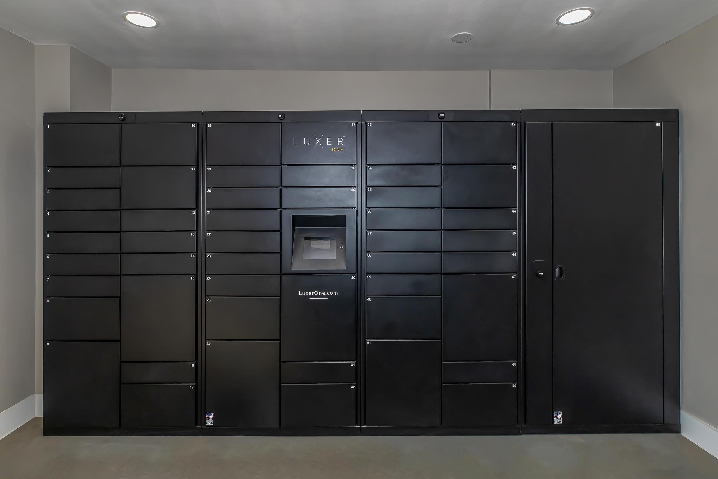 A large black locker system mounted on a wall, featuring multiple compartments for package storage. The center displays a digital interface, likely for managing deliveries. The overall design is modern and sleek, set against a neutral-colored wall in a well-lit indoor space.