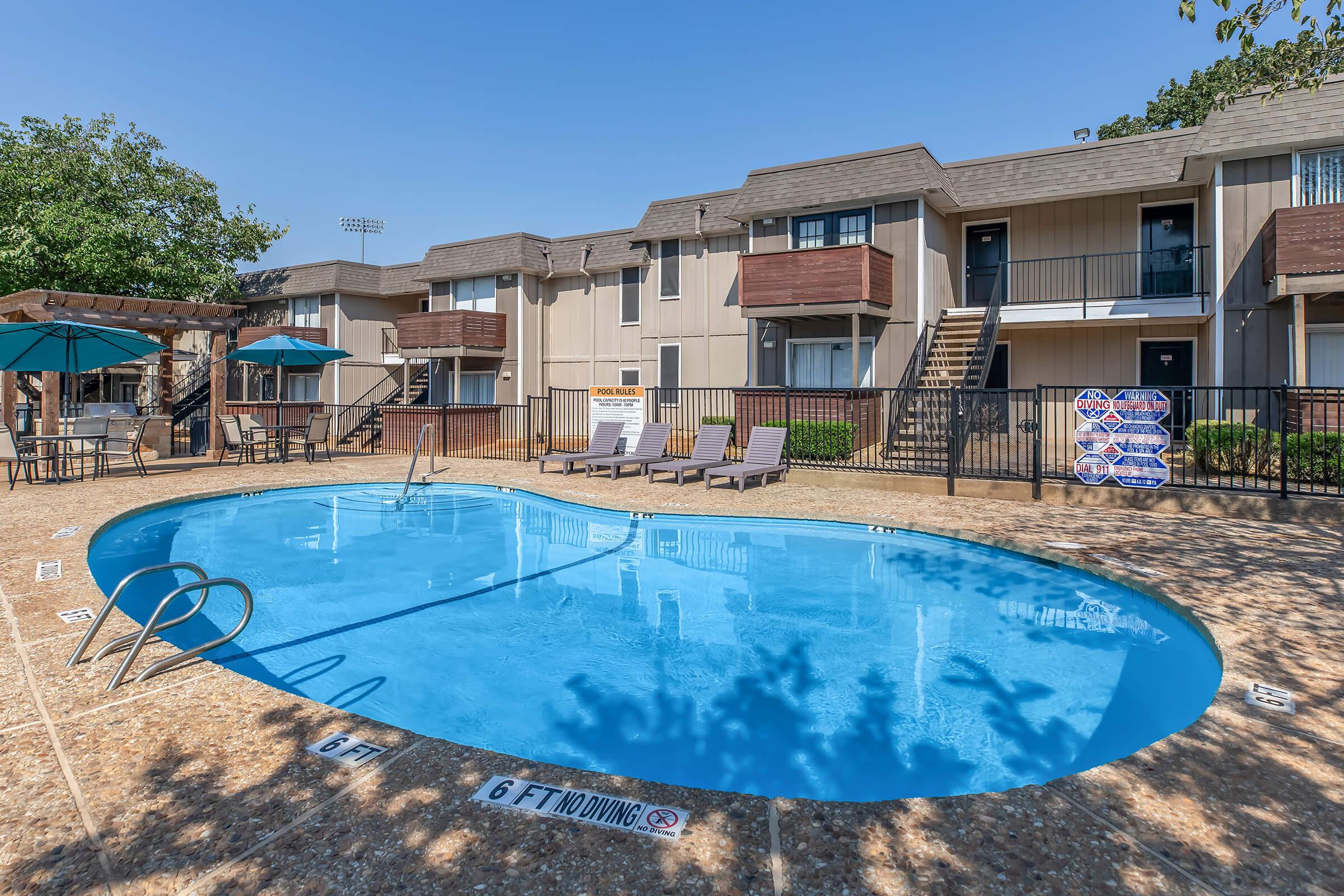 A clear blue swimming pool surrounded by lounge chairs, with an apartment building in the background. There are umbrellas for shade, and signs indicating the pool rules. The sky is sunny and blue, creating a relaxed outdoor atmosphere.