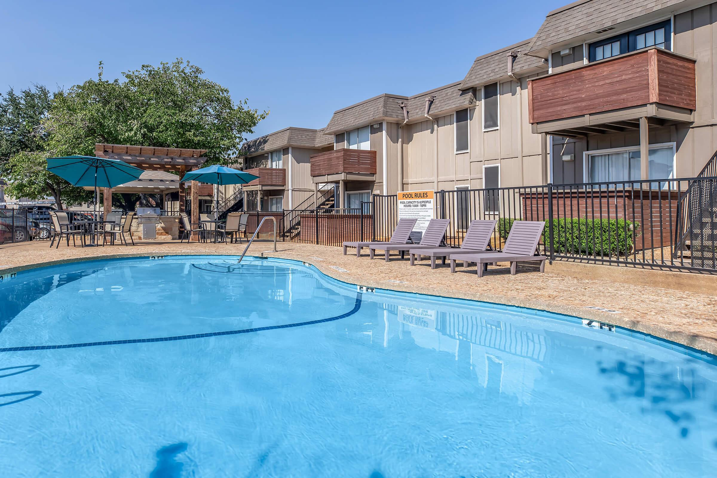 A swimming pool with clear blue water and sun loungers around it, surrounded by a fenced area. In the background, there are apartment buildings with wooden balconies. Sunny weather with a few trees providing shade. Umbrellas over some seating areas near the pool.