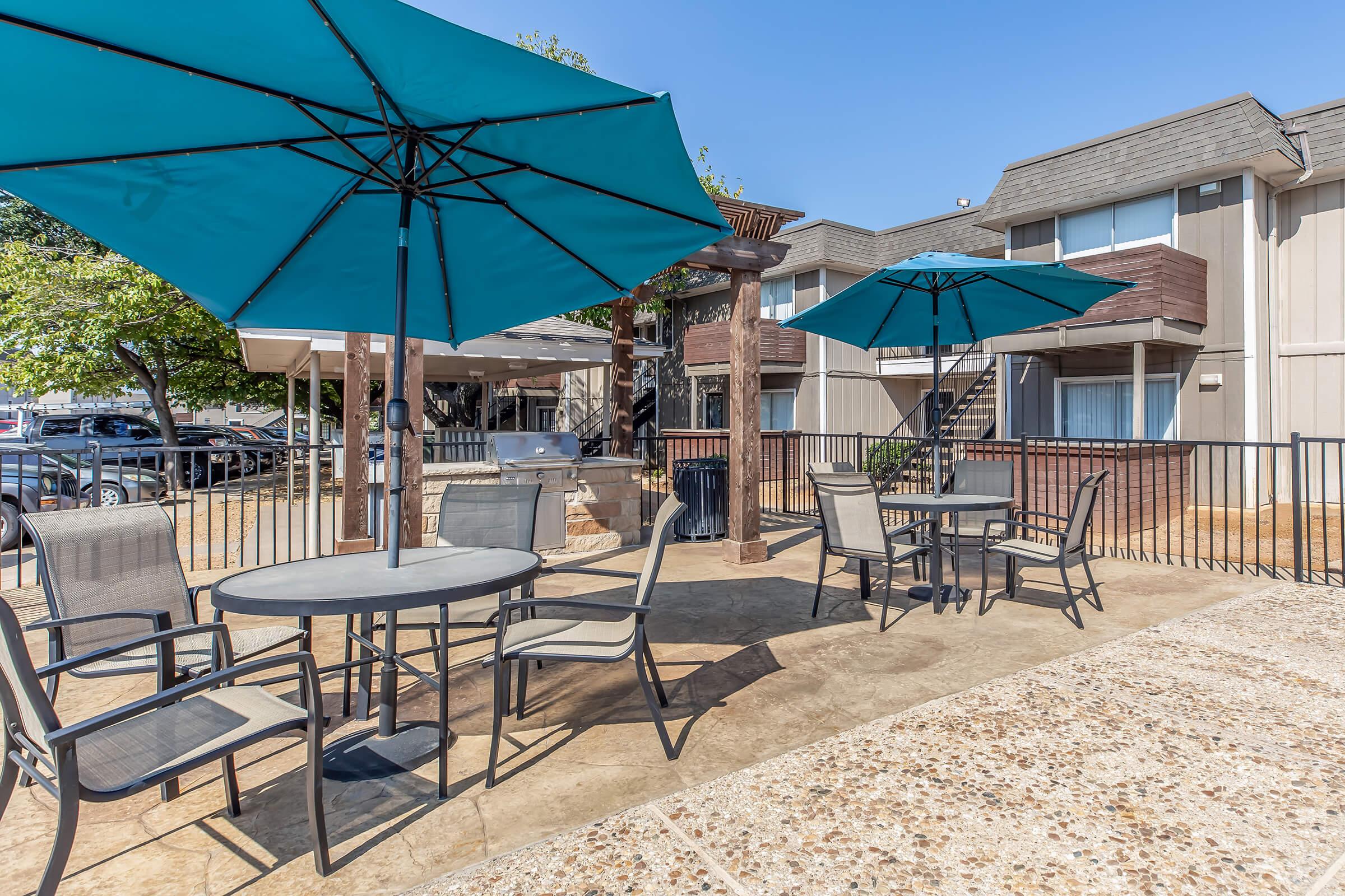 A patio area with several tables and chairs under large blue umbrellas. The setting includes a grill and landscaped greenery, with a backdrop of apartment buildings and a clear blue sky. The concrete surface is textured, creating a cozy outdoor dining space.