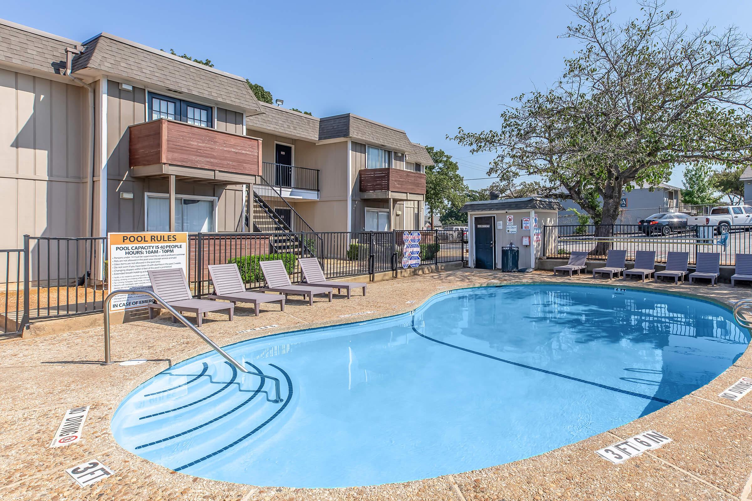 A sunny outdoor swimming pool area with clear blue water, surrounded by lounge chairs. In the background, there are two-story apartment buildings. A sign detailing pool rules is visible. Trees provide shade near the edge of the pool. The overall scene is inviting and well-maintained.