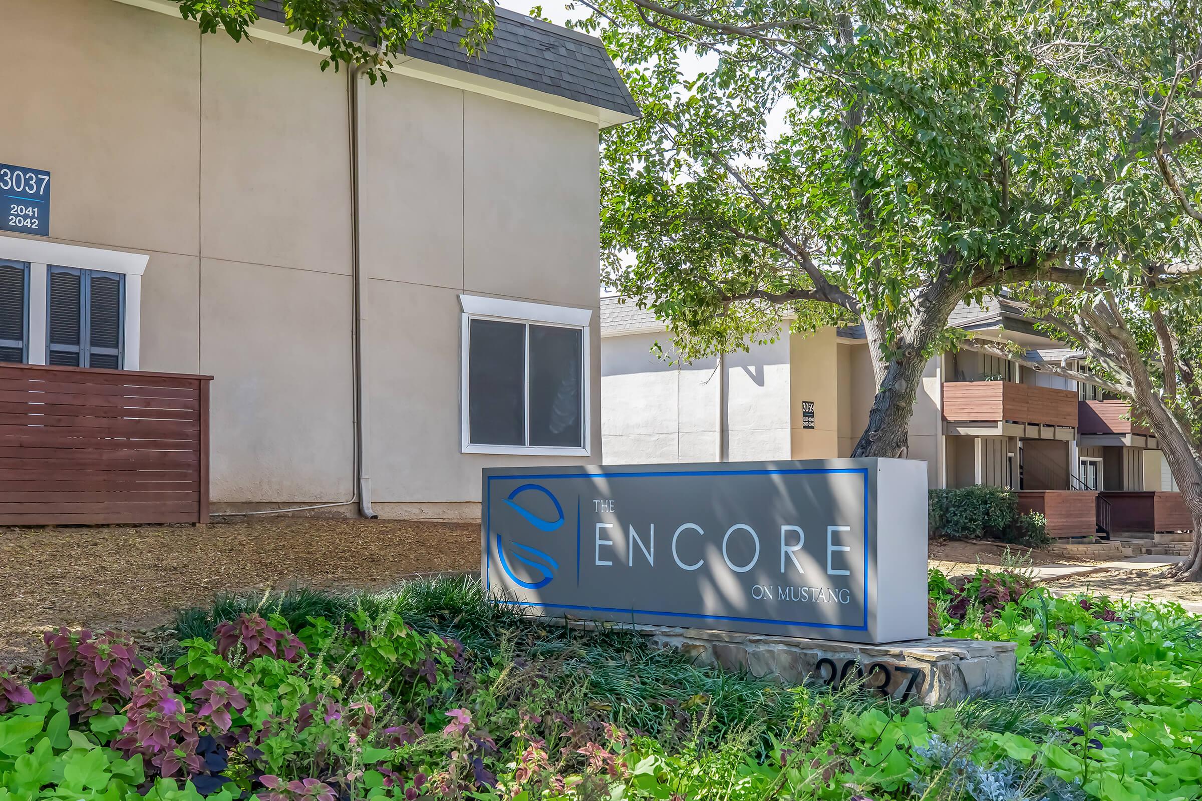 Sign for "The Encore on Mustang" at an apartment community, with the building in the background surrounded by greenery and landscaping. The sign is prominently displayed near the entrance, indicating the name and location of the residential property.