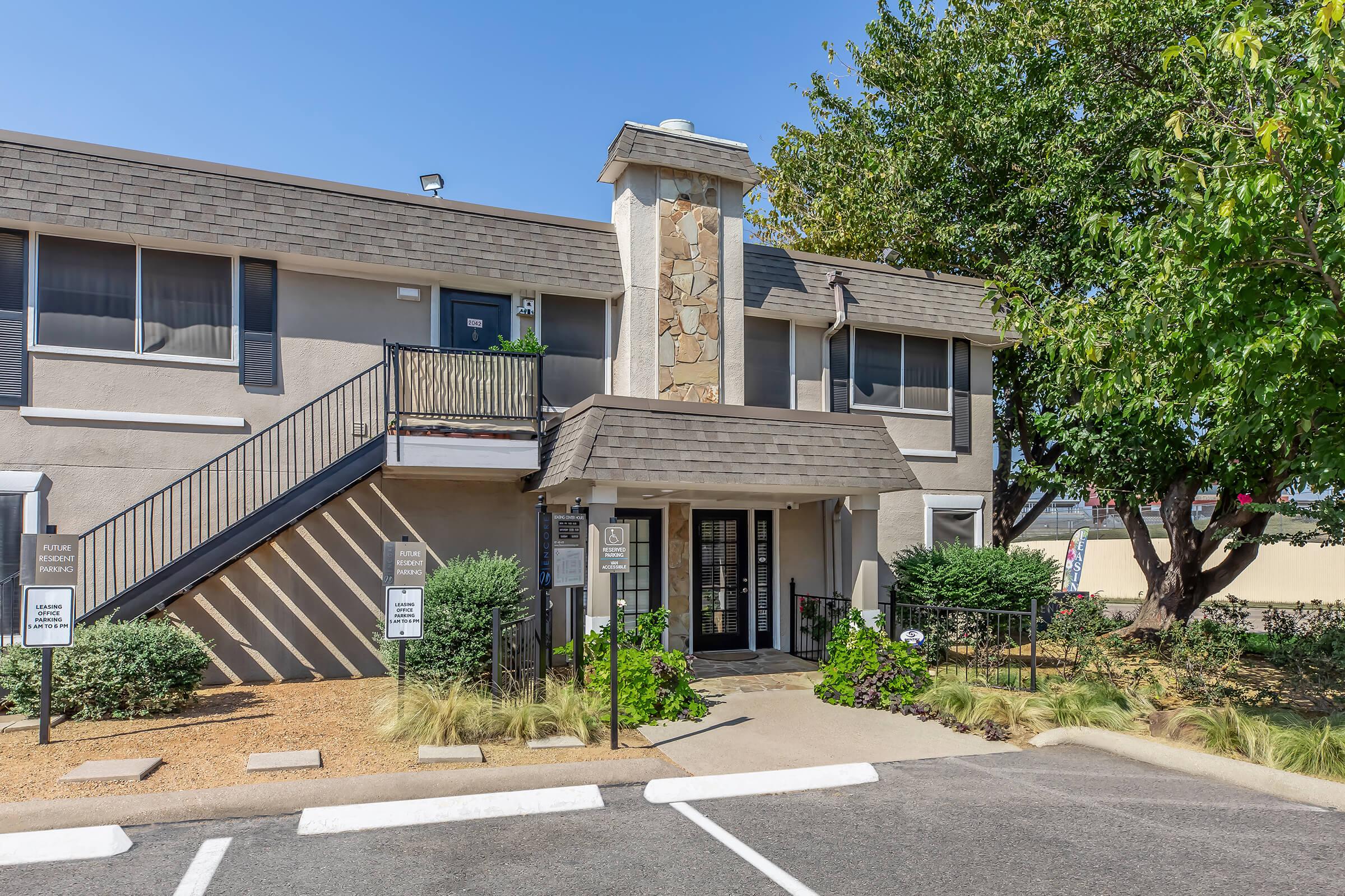 A two-story residential building with a stone chimney and exterior stairs leading to the upper floor. The entrance features a double door, framed by landscaped shrubbery and decorative plants. Nearby, there are parking spaces marked in front of the building, under a clear blue sky.