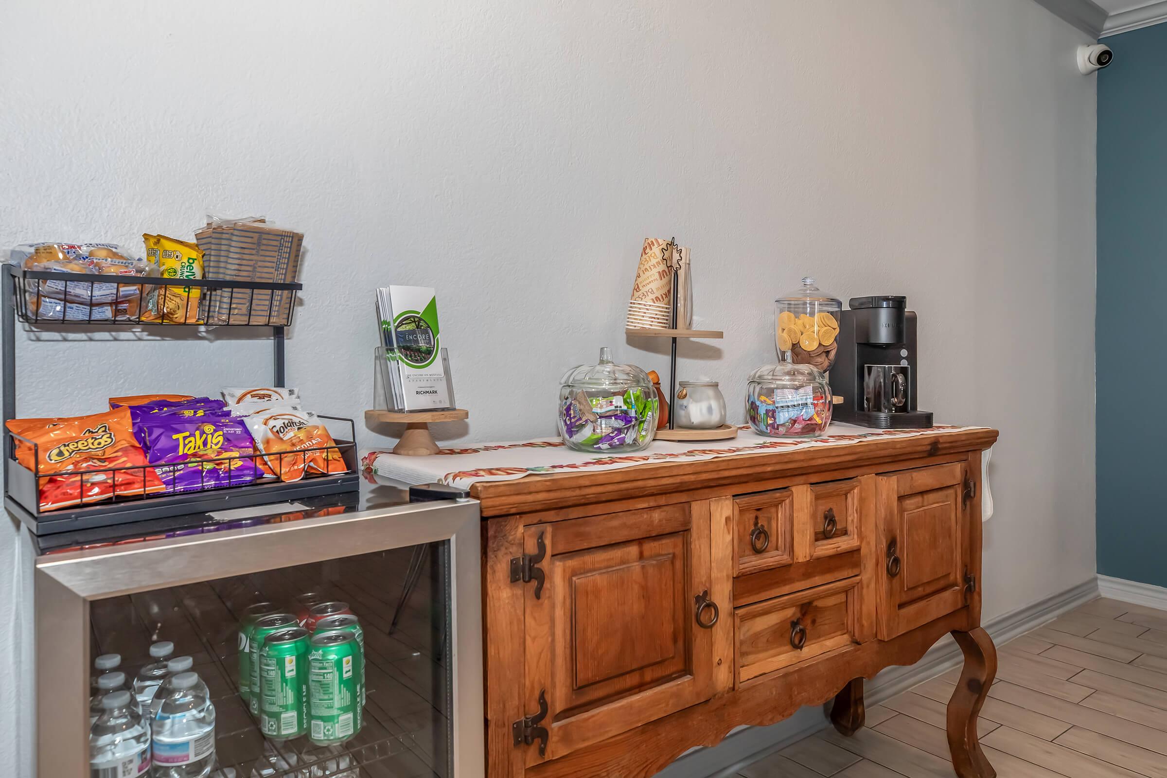 A wooden sideboard displaying various snacks, candy, and drinks. A small beverage cooler is visible underneath, stocked with cans. There's a coffee machine and glass containers filled with cookies and treats, alongside a promotional sign. The setting has a clean, modern look with light-colored walls.