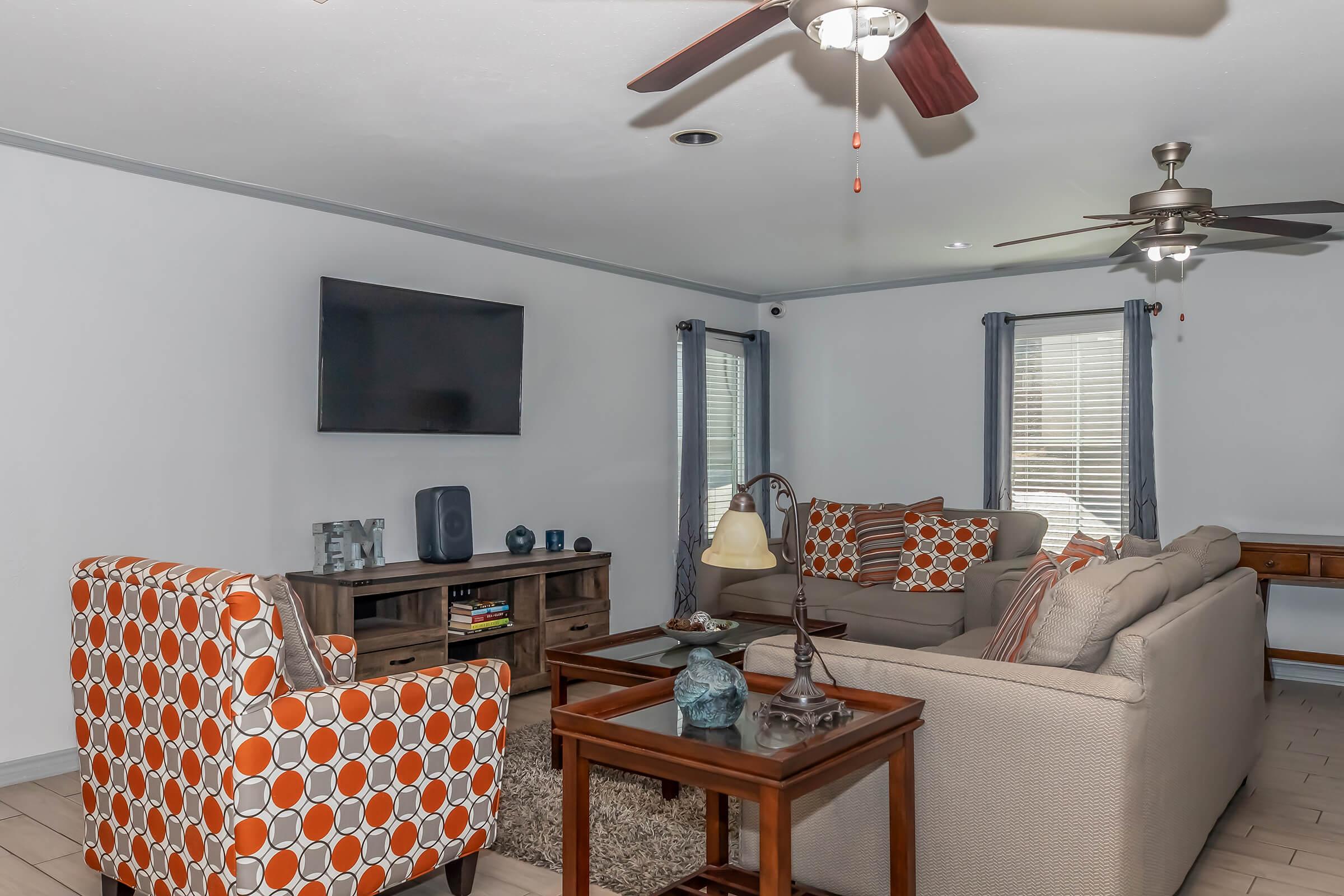 A cozy living room featuring a beige sofa with orange and gray pillows, an orange patterned armchair, a wooden coffee table with decorative items, and a TV mounted on the wall. Natural light streams through two windows with gray curtains, and a ceiling fan adds to the comfortable atmosphere.