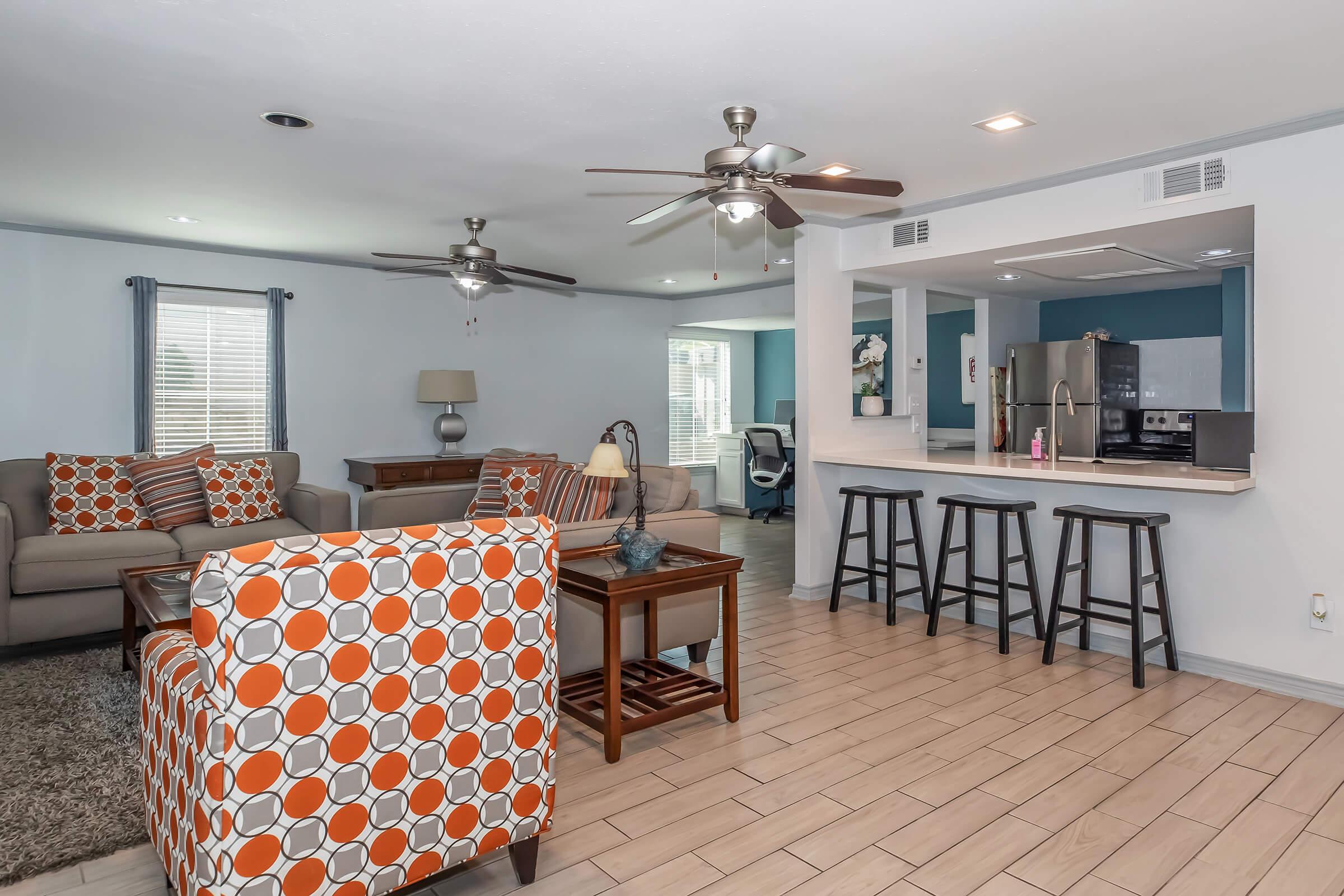 A bright and airy living area featuring a comfortable seating arrangement with patterned orange and gray chairs, a tan sofa, and a coffee table. In the background, there's a kitchen area with modern appliances and bar stools. The walls are painted light blue, creating a fresh and inviting atmosphere.