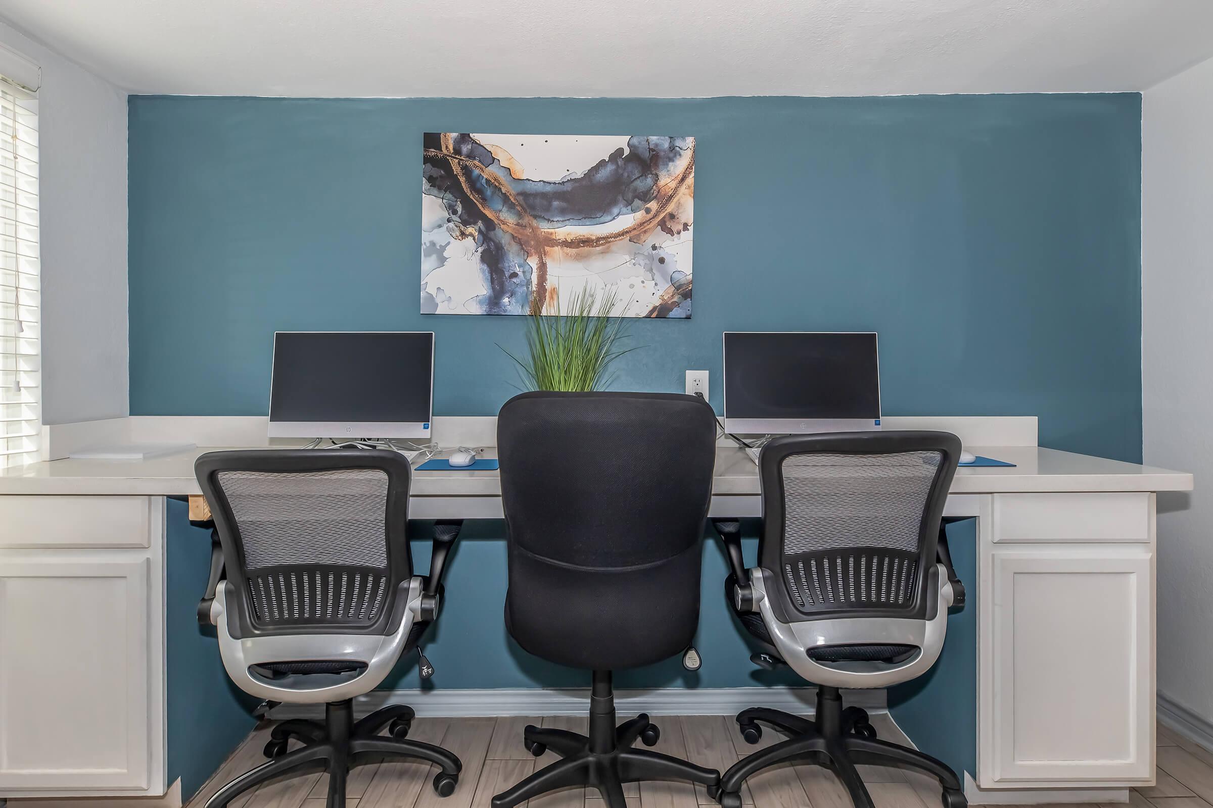 A modern home office featuring two ergonomic chairs positioned at a light-colored desk with two monitors. A decorative abstract painting in blue and gold hangs on the wall, and a small green plant adds a touch of greenery to the workspace. The wall is painted in a calming teal color.