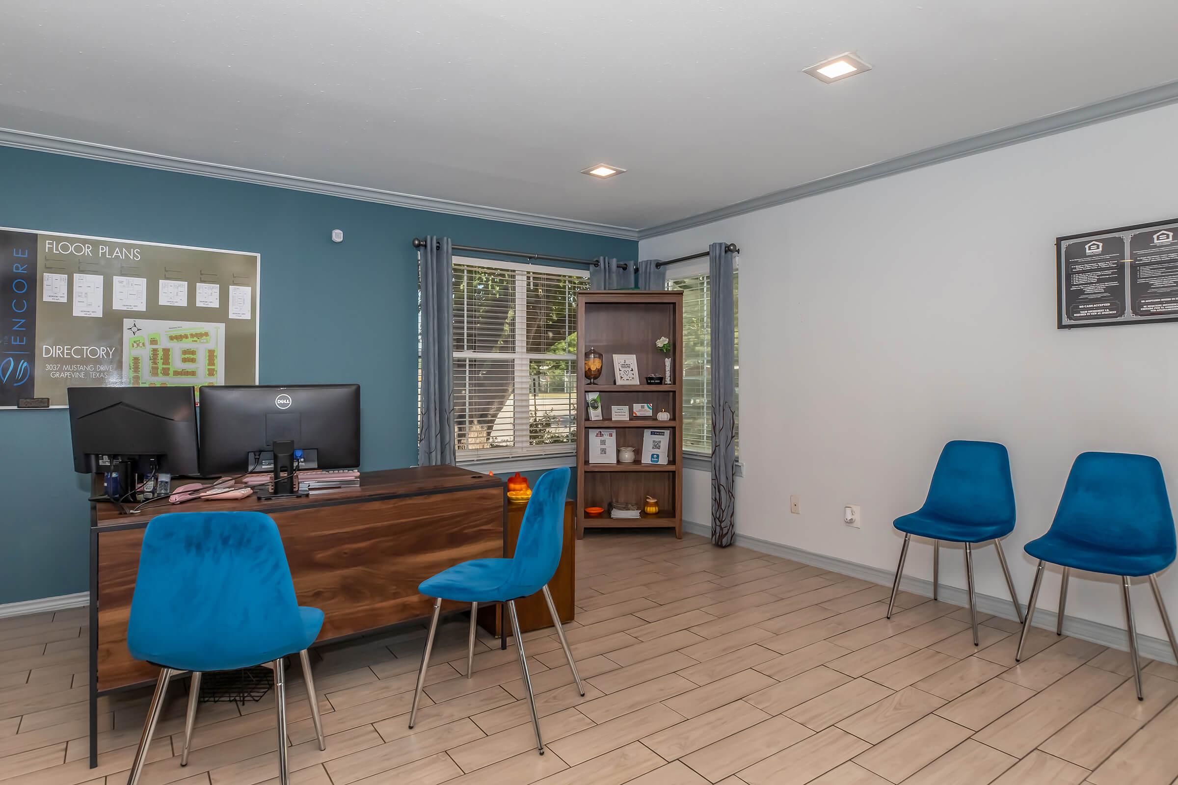 A modern office reception area featuring a desk with two monitors, brochures, and a decorative shelf. Blue walls complement light tile flooring. In the foreground, there are four stylish blue chairs arranged around the desk, creating a welcoming atmosphere for visitors. Natural light comes in through large windows.