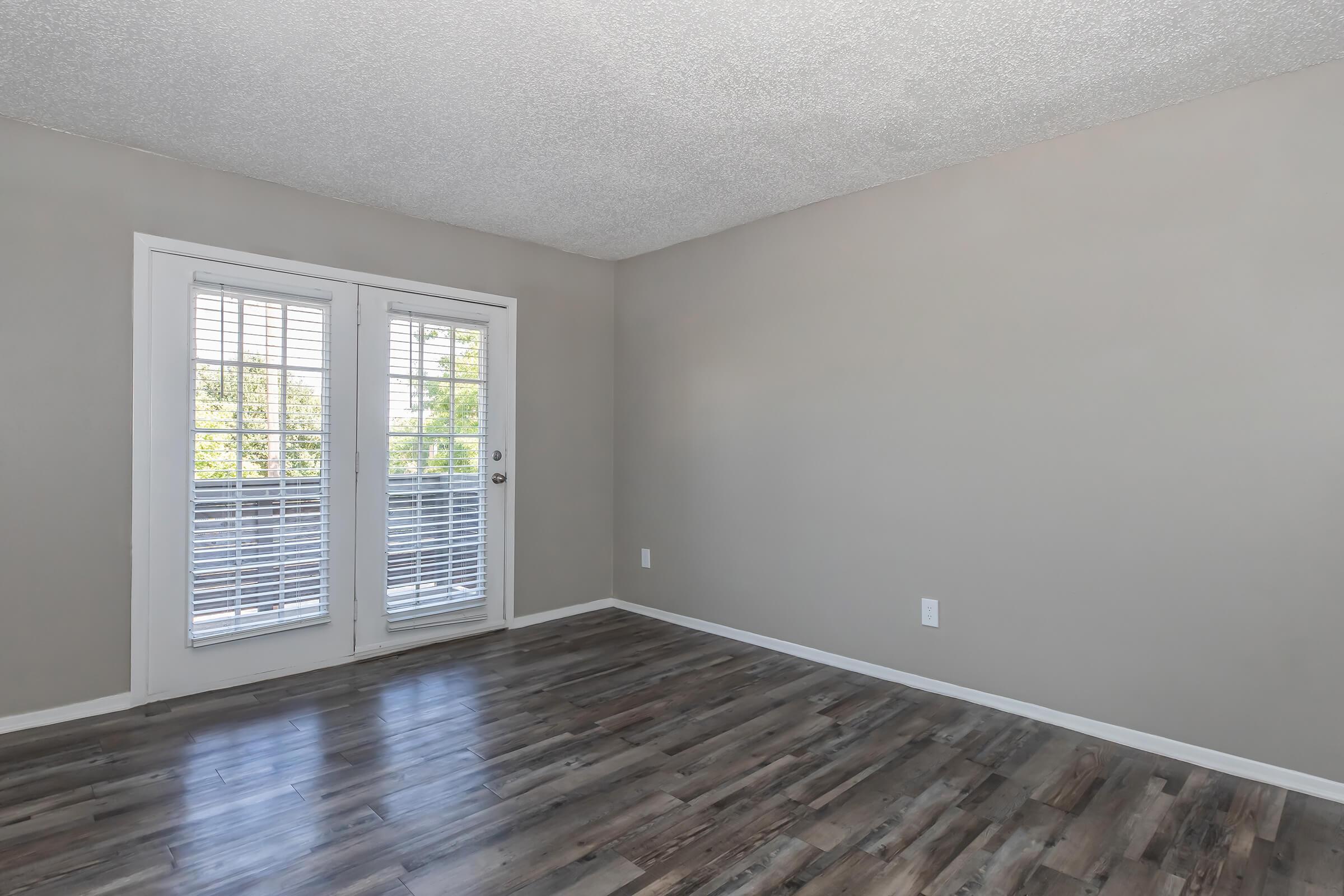 Empty room with light gray walls and a textured ceiling. Features two sliding glass doors with white blinds that open to a balcony. The floor has dark wooden laminate, and there are no furnishings or decorations, creating a spacious and minimalist appearance.