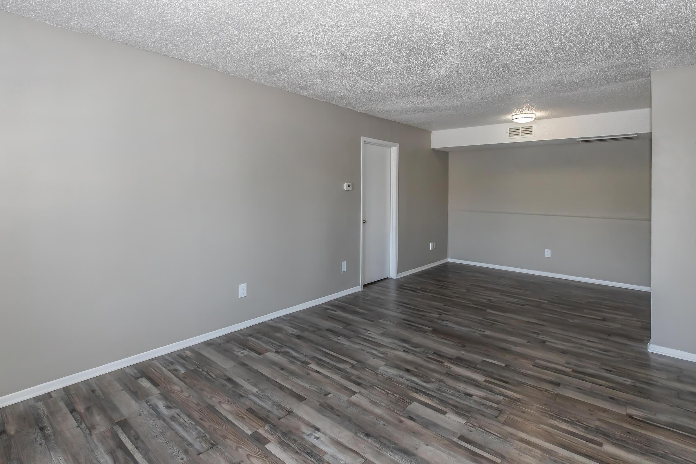 A spacious, empty room with light gray walls and a textured ceiling. The floor is covered in wood-like laminate, and there is a white door on the left side. The room has a ceiling light fixture and no furniture, creating a clean, open atmosphere suitable for decorating.