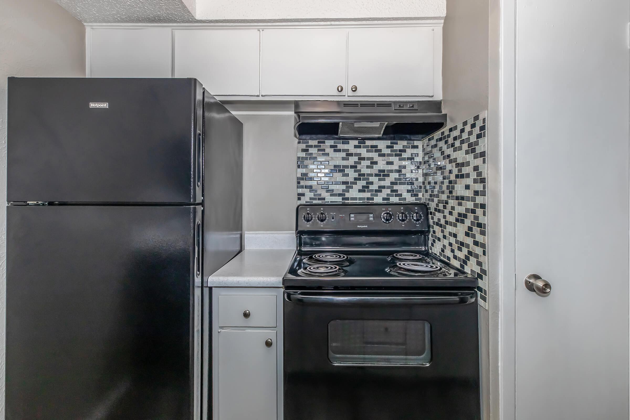 A modern kitchen featuring a black refrigerator and an electric stove with a black finish. The stove has four burners and a hood vent above it. The backsplash consists of small mosaic tiles in shades of gray and black, and there are white cabinets above the appliances, along with a light-colored countertop.