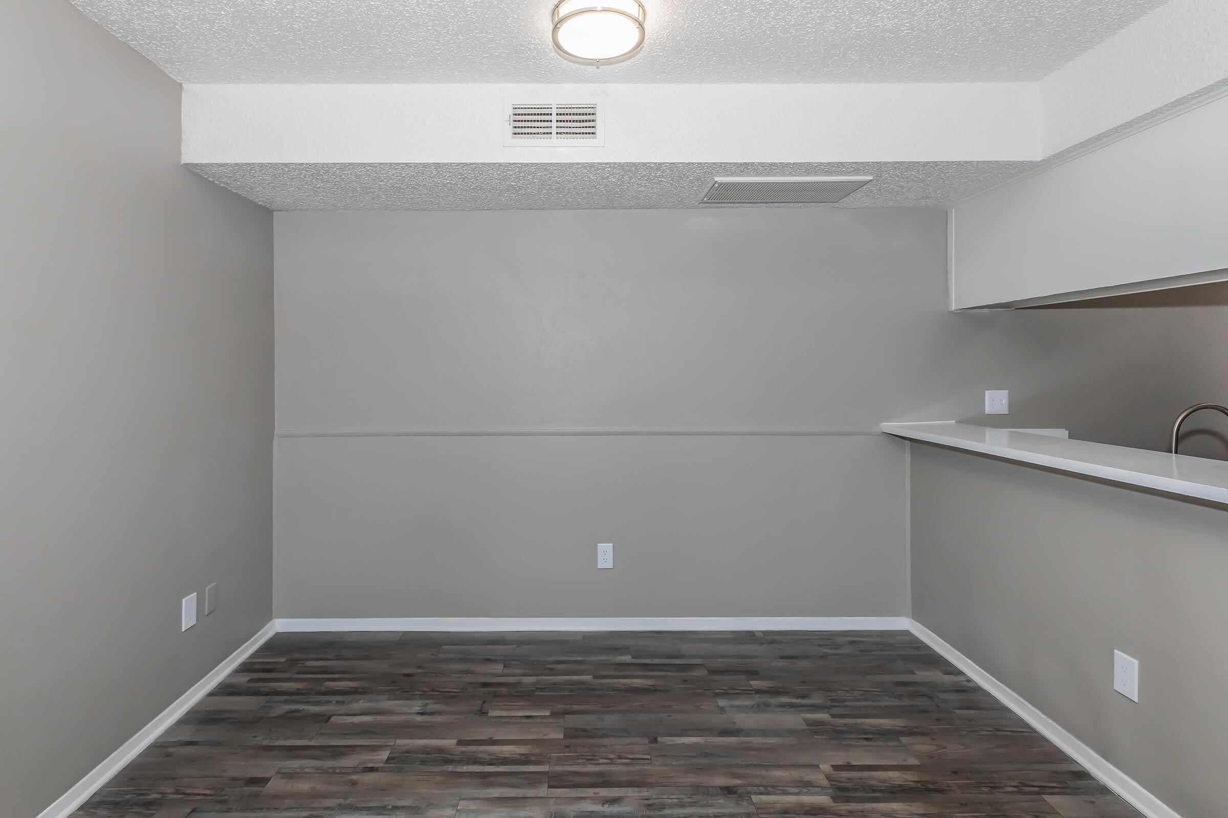 A spacious, empty room with light gray walls and a darker wood-like floor. A small kitchen area is visible on the right with a bar-style counter. The ceiling features a circular light fixture, and there are no furnishings or decorations present. The overall atmosphere feels clean and modern.