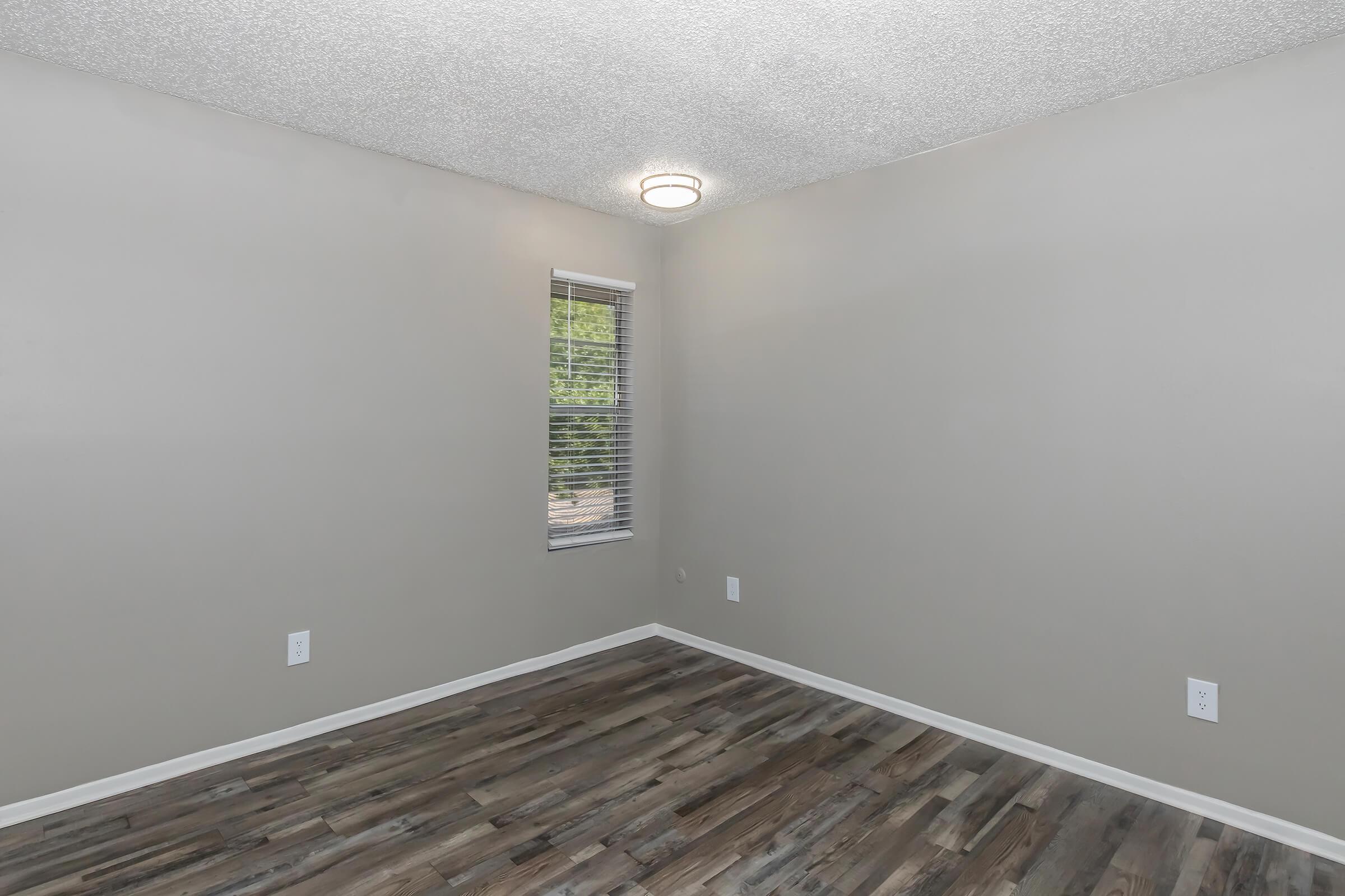 A sparsely furnished room with light gray walls and a textured ceiling. It features a ceiling light fixture, a small window with blinds, and wooden-like flooring. The space appears clean and empty, suitable for various uses or decoration.