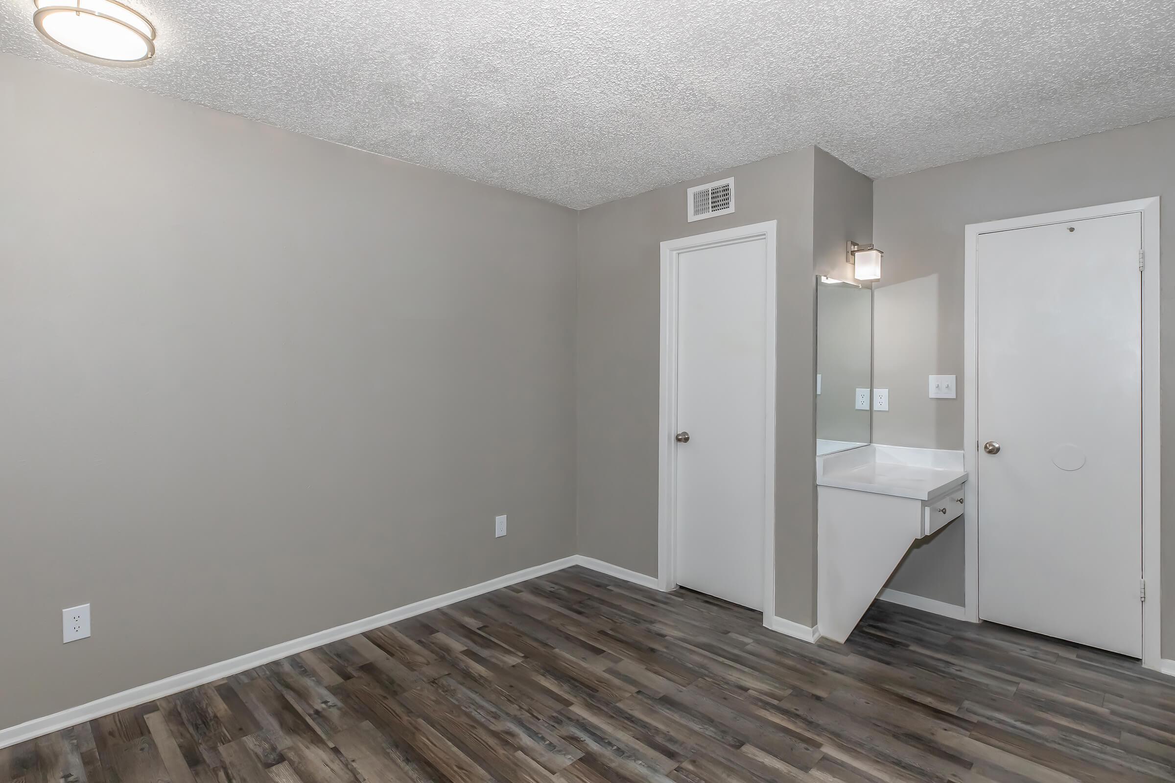 A room with gray walls and a textured ceiling, featuring a light fixture, a large mirror above a white vanity, and a door leading to another room. The floor is covered in laminate wood, creating a clean and modern look.