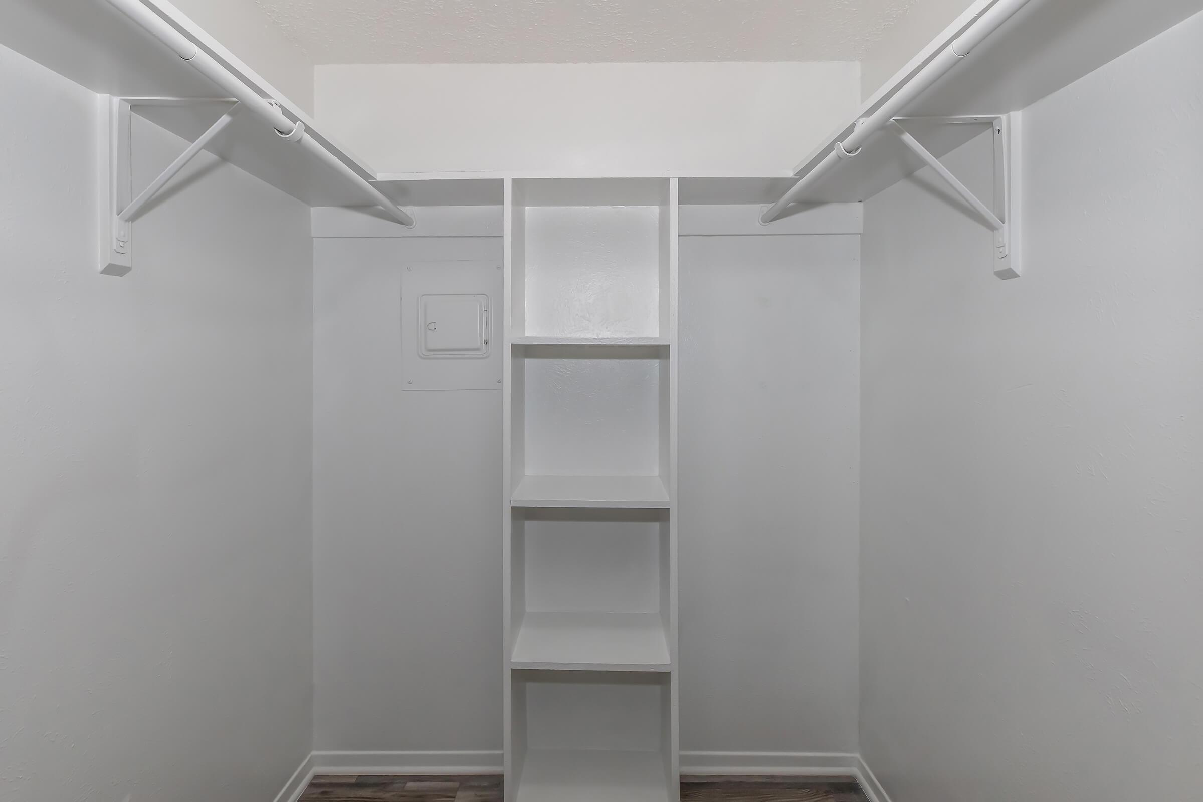 A spacious, empty closet featuring two horizontal hanging rods and a central shelving unit with five shelves. The walls are painted white, and the floor has a wooden appearance. The overall design is minimalistic and clean, primarily focusing on functionality.