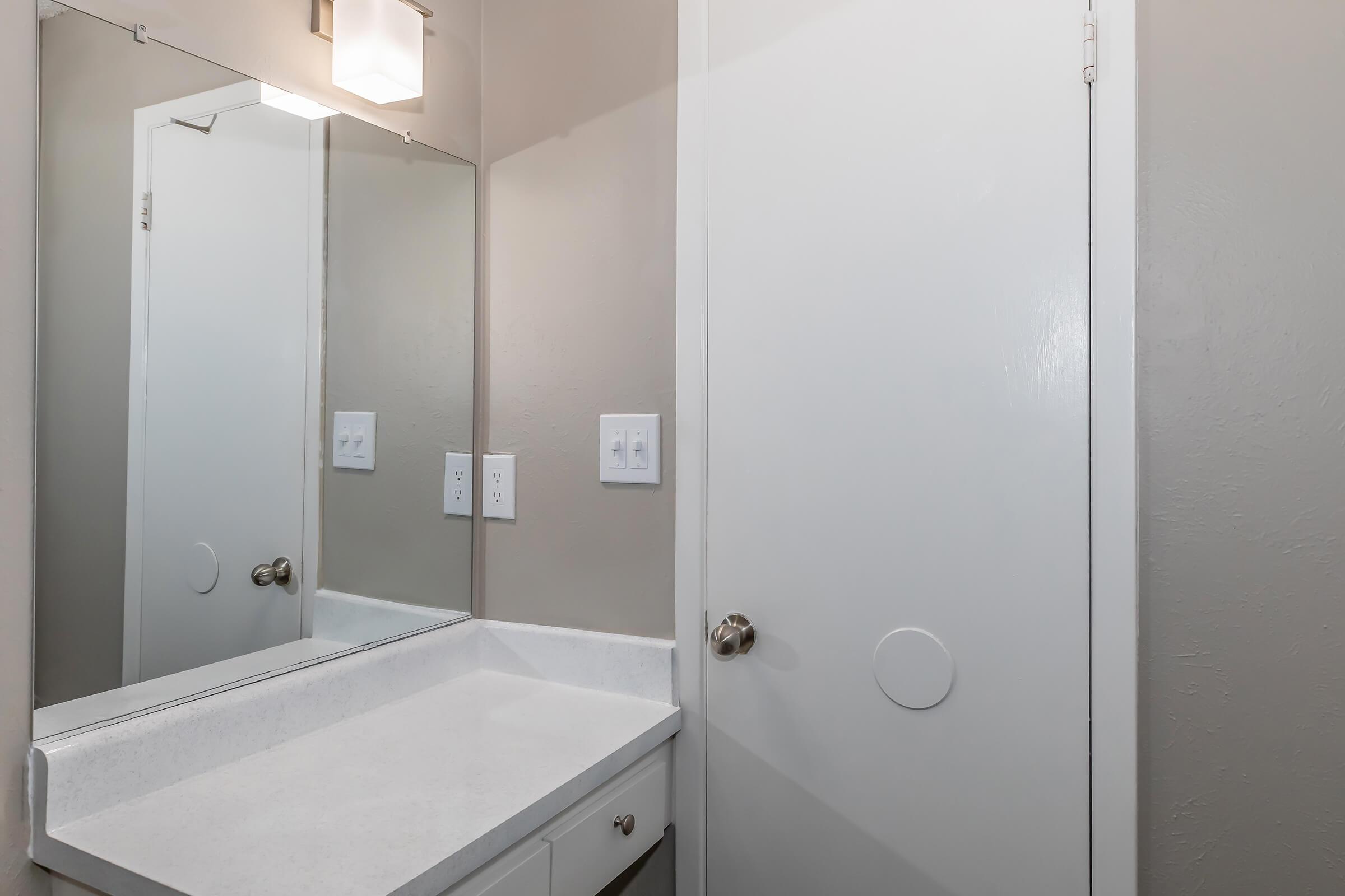 A small bathroom with a simple white counter, a rectangular mirror above it, and light gray walls. A closed door is visible on the right side, and there are two light switches on the wall next to the mirror. The overall design is clean and minimalistic.