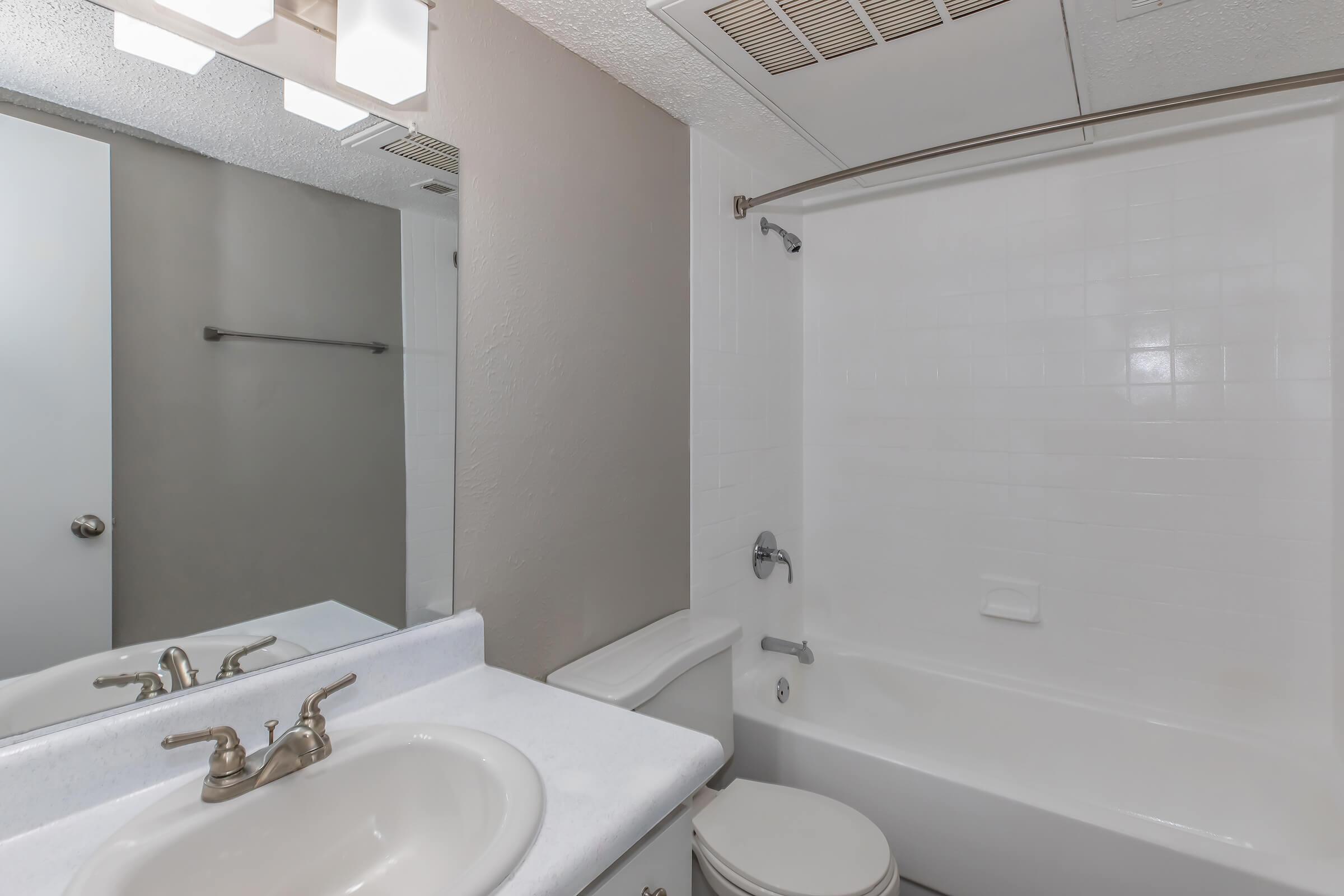 A clean and simple bathroom featuring a white bathtub with a shower, a sink with a countertop, and a large mirror above the sink. The walls are painted in neutral colors, and there is a towel bar on one side. The space is well-lit with overhead lighting.