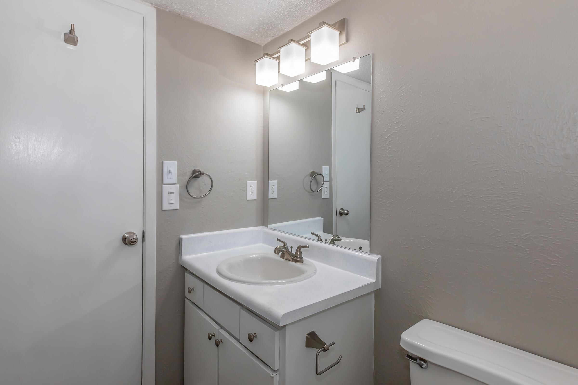 A modern bathroom featuring a white sink with a gold faucet on a white countertop, a large mirror, and a light fixture above. The walls are painted in a neutral gray color, and there is a white toilet to the right. The door is partially visible, showing a silver doorknob.