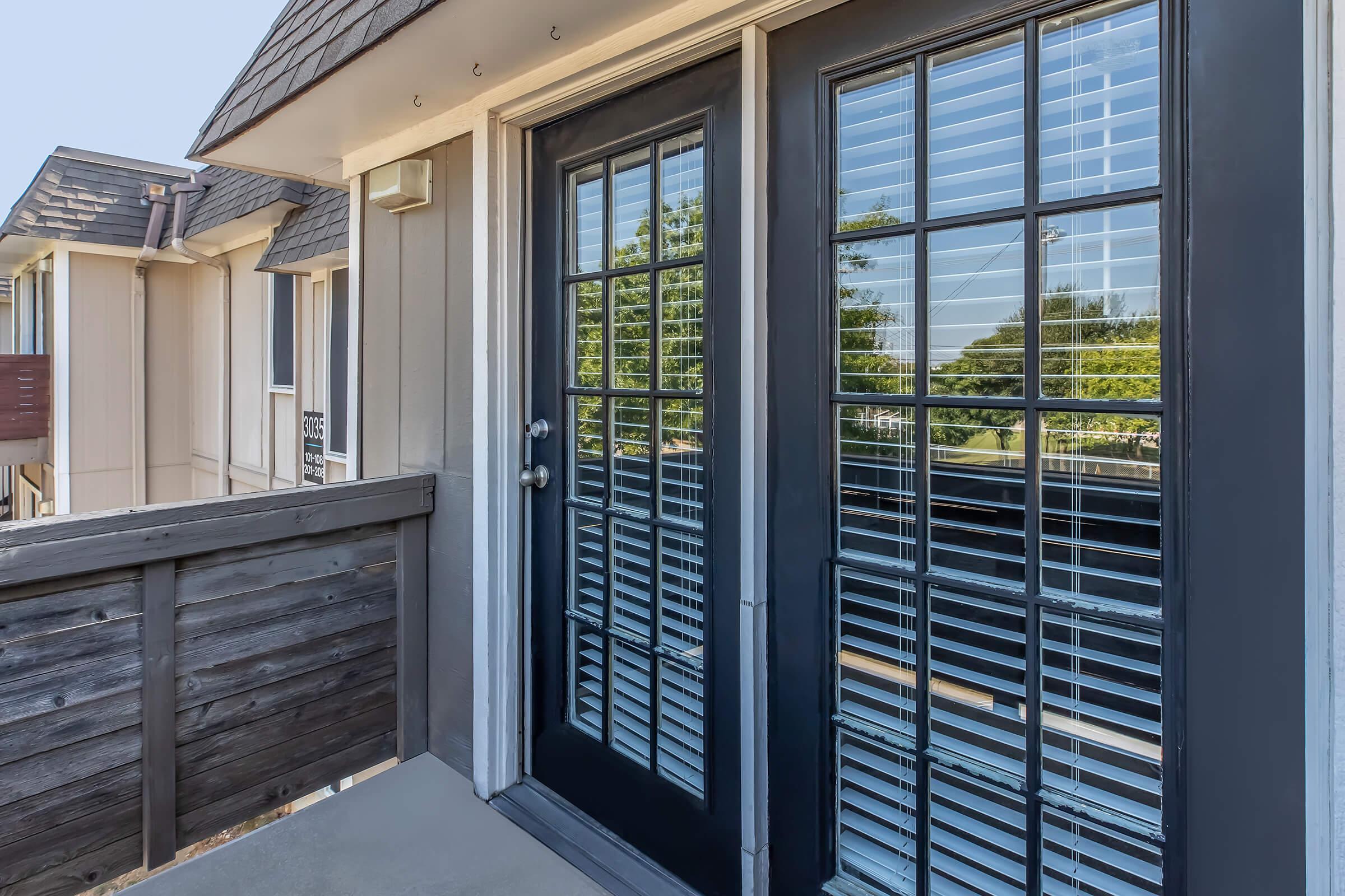 A set of double doors with black frames and horizontal blinds, leading to a balcony or patio. The doors are partially open, revealing a view of green trees outside. The exterior features muted colors, with wood paneling visible on a nearby railing. Clear blue skies are visible above.