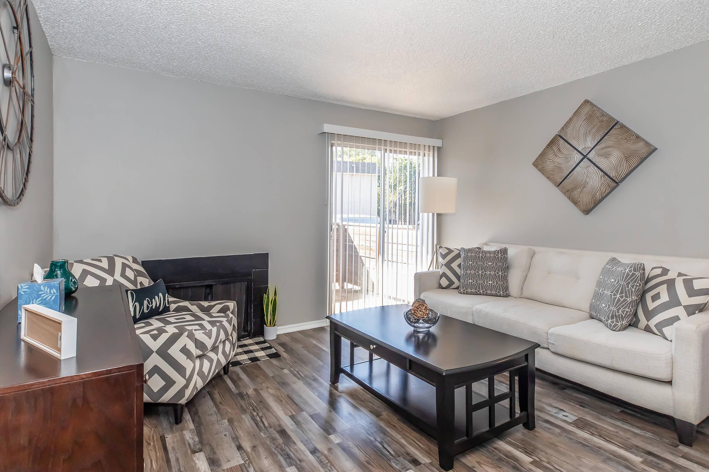 A modern living room featuring a light gray wall, a comfortable beige couch with decorative pillows, a patterned chair, a dark wooden coffee table, and a stylish wall clock. Natural light streams in from sliding glass doors, and the floor has a wood-like finish, creating a welcoming atmosphere.