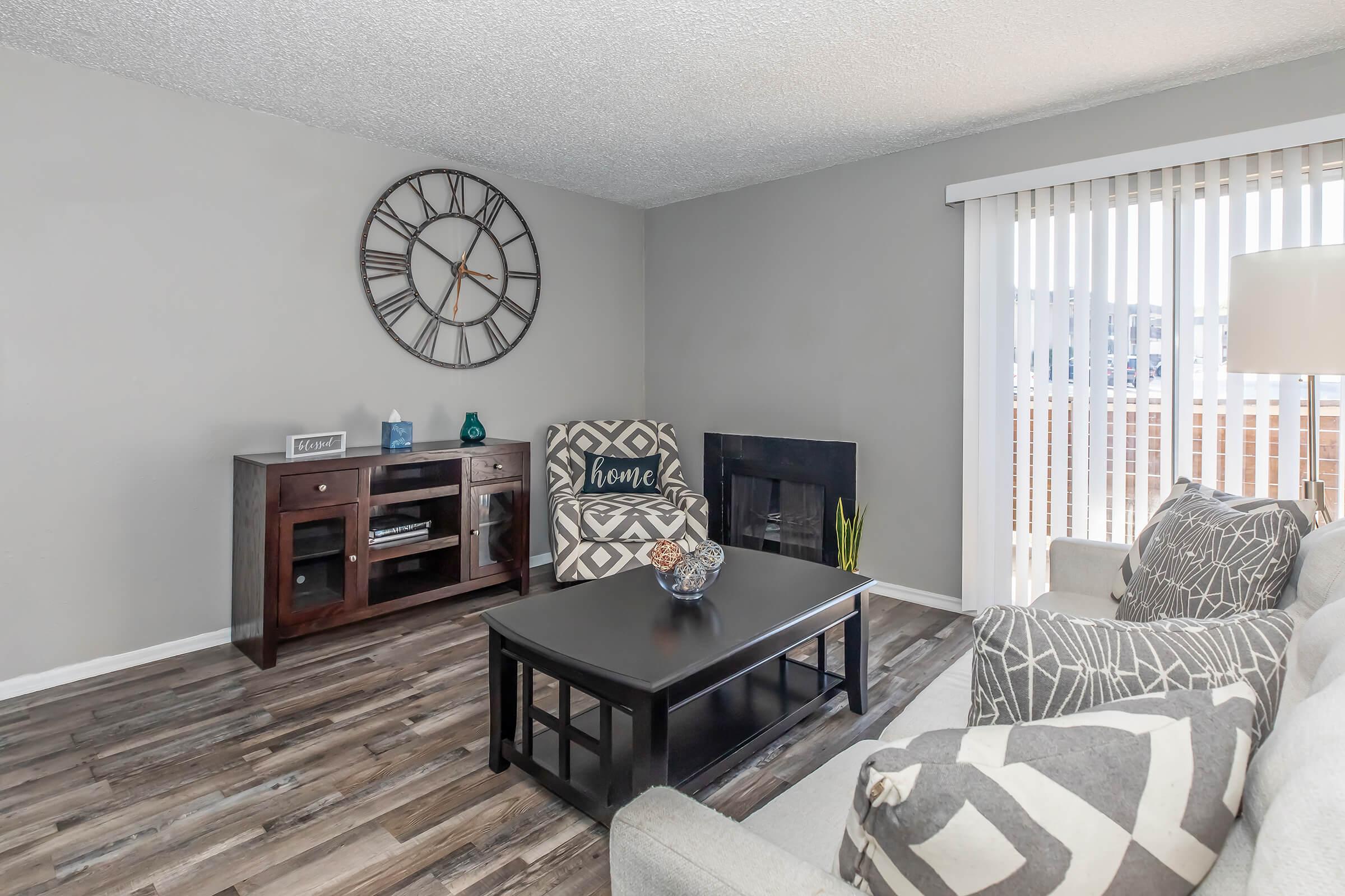 A modern living room featuring a light gray color scheme, with a large wall clock, a dark wooden TV stand, and a black fireplace. There are two patterned armchairs and a cozy sofa, complemented by a coffee table with decorative items. Vertical blinds cover a window, allowing natural light to enter.