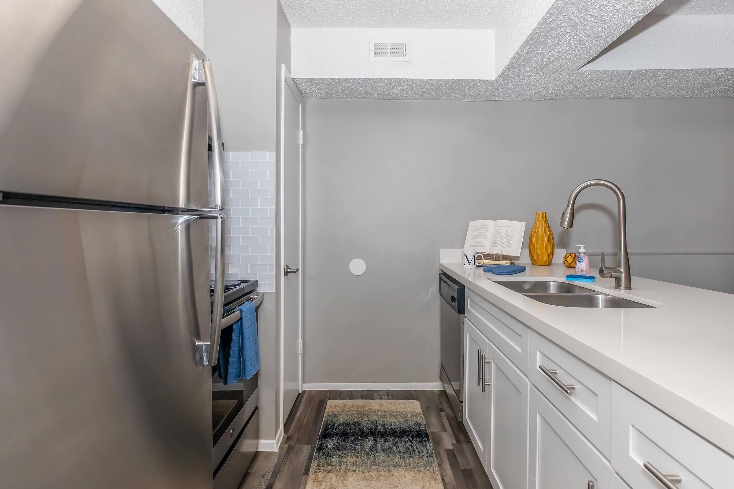 A modern kitchen featuring stainless steel appliances, including a fridge and stove. The countertop is equipped with a sink and minimal decor, including a yellow vase and a cookbook. The walls are painted in neutral tones, and there's a rug on the floor, enhancing the contemporary design of the space.