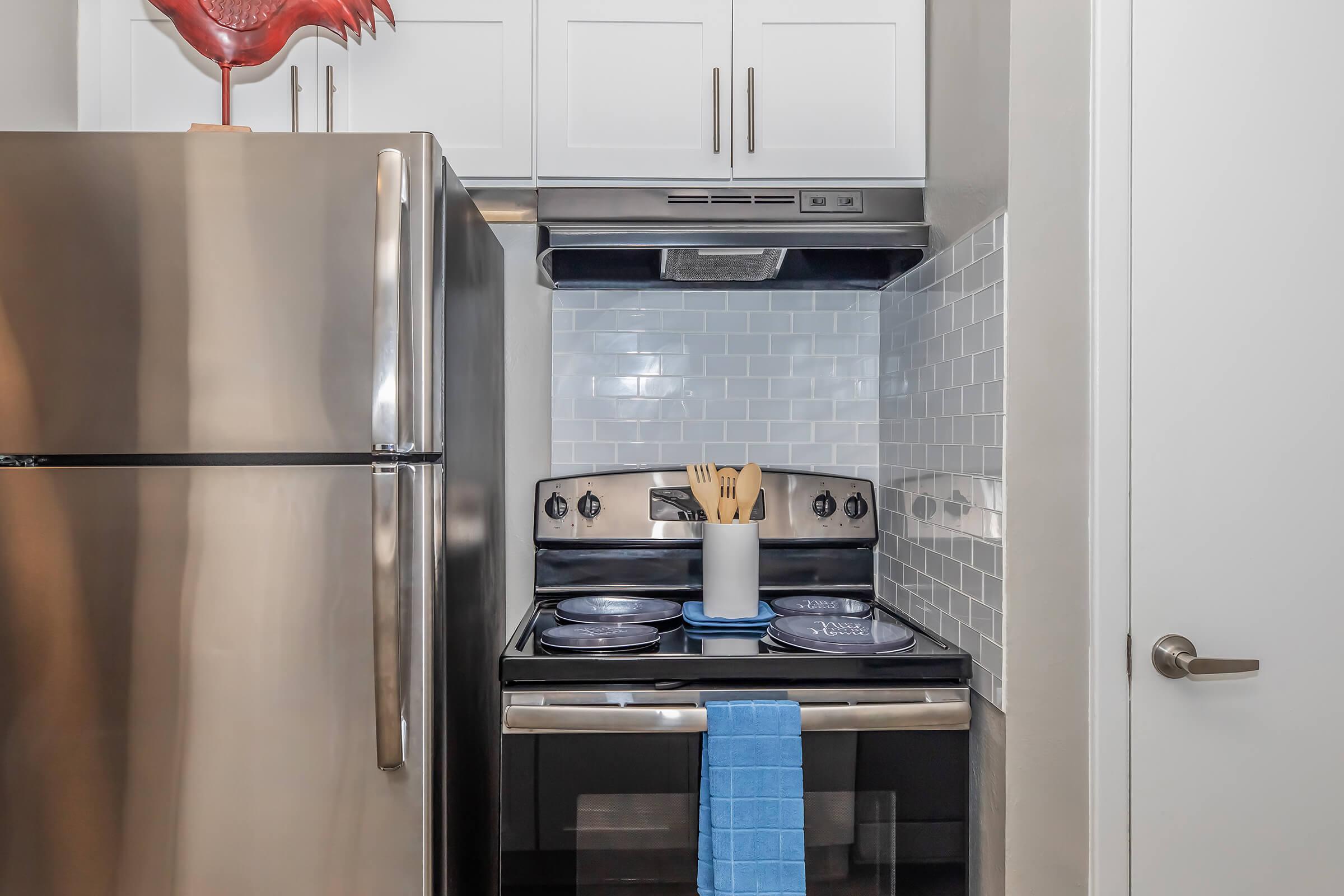 A modern kitchen featuring a stainless steel refrigerator and an electric stove with a silver hood. The stove has a container of wooden cooking utensils, and a blue towel is hanging from the oven handle. The backsplash is made of light blue tiles, adding a pop of color to the neutral decor.