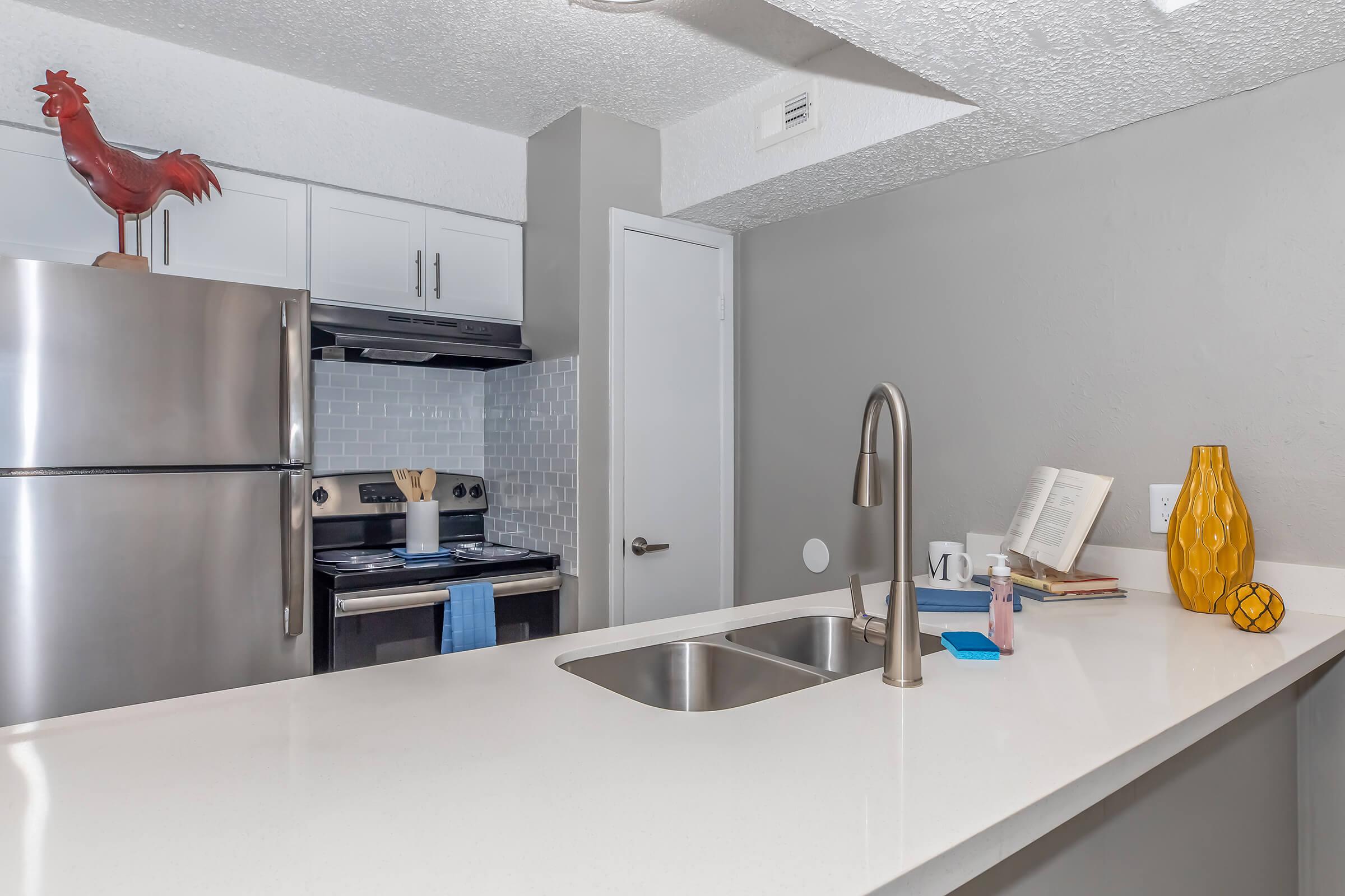 A modern kitchen featuring stainless steel appliances, a white countertop with a double sink, and light gray walls. Decorative items include a yellow vase, a book on the counter, and a rooster figurine on top of the cabinets. The space is bright and well-organized, conveying a clean and inviting atmosphere.