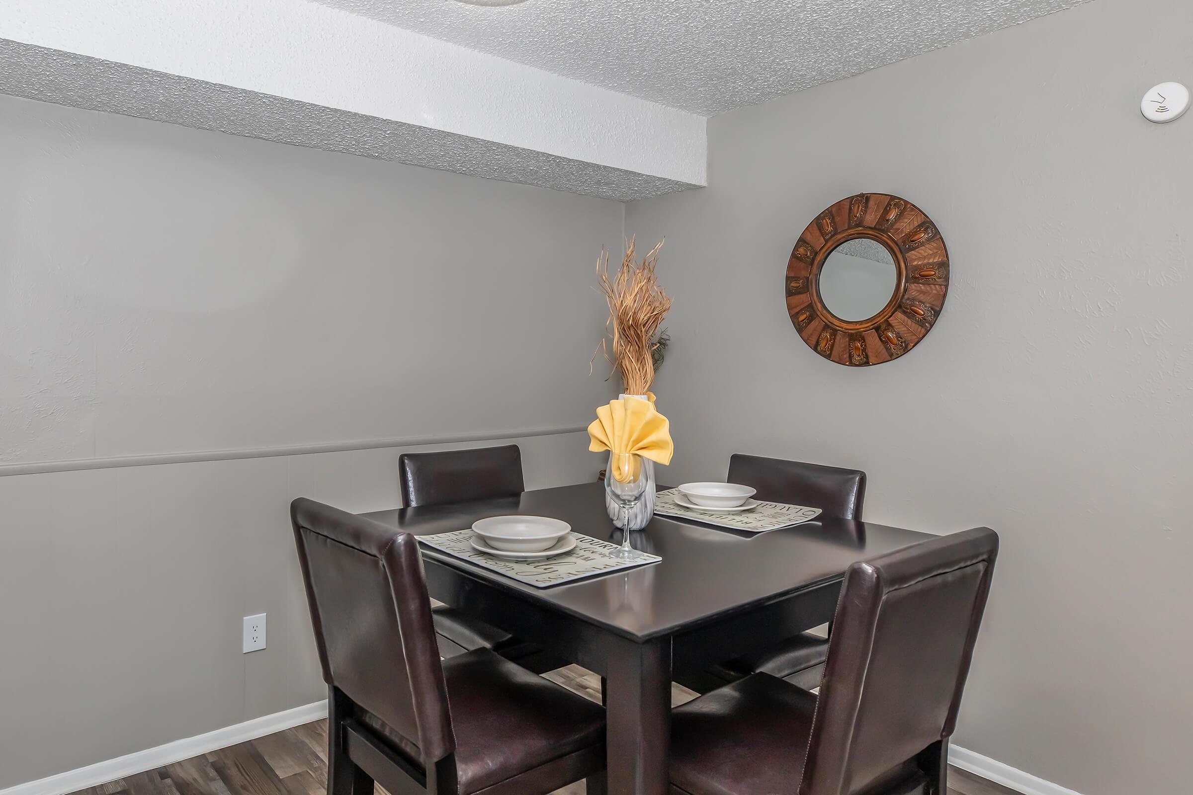 A cozy dining area featuring a square black table with four brown leather chairs. The table is set with plates and a decorative vase holding a yellow flower. A round mirror with a wooden frame hangs on the wall, and dried decorative grass adds a natural touch to the decor. The walls are painted a soft gray.