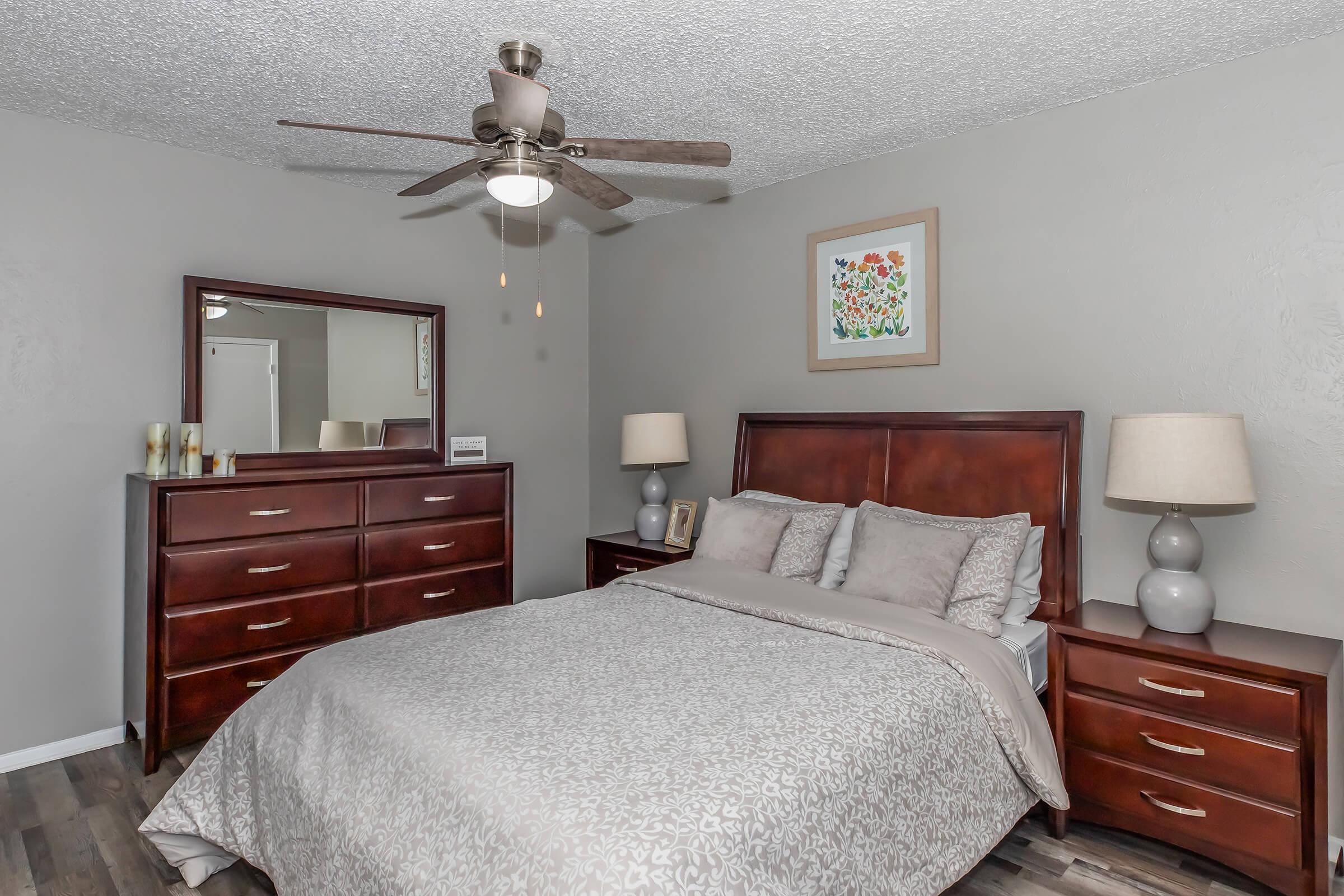 A cozy bedroom featuring a wooden bed with soft gray bedding, two bedside lamps, and a matching dresser. A large mirror above the dresser enhances the space, while a colorful framed artwork adds a touch of decor. The room has a ceiling fan and a neutral-colored wall, creating a warm and inviting atmosphere.