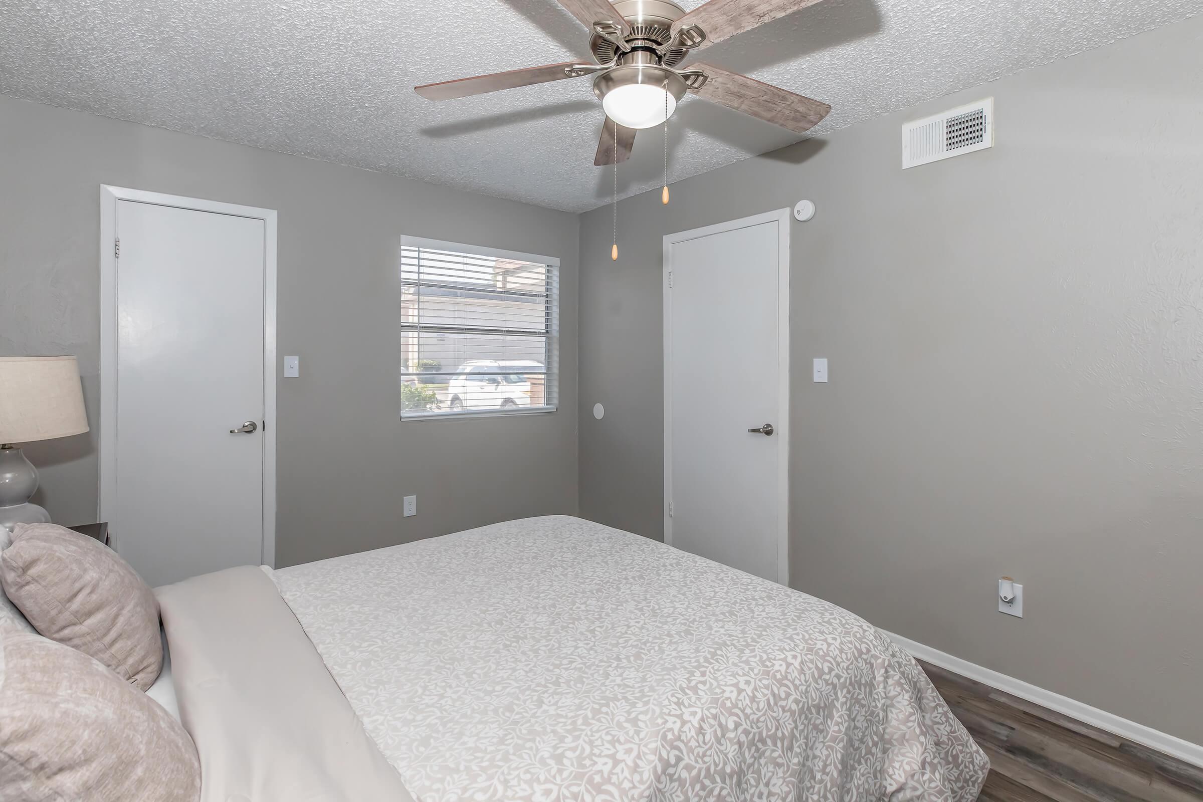 A cozy bedroom featuring a queen-sized bed with light bedding. Two doors are visible on the walls, one leading outside and the other likely to a closet. A window allows natural light in, and a ceiling fan is mounted above, enhancing the room's comfort. The walls are painted in a neutral gray tone.